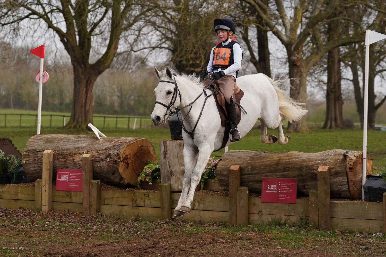 A young rider in riding gear and a helmet jumping over a log obstacle on a white horse during a cross-country equestrian event in an outdoor setting with trees in the background.