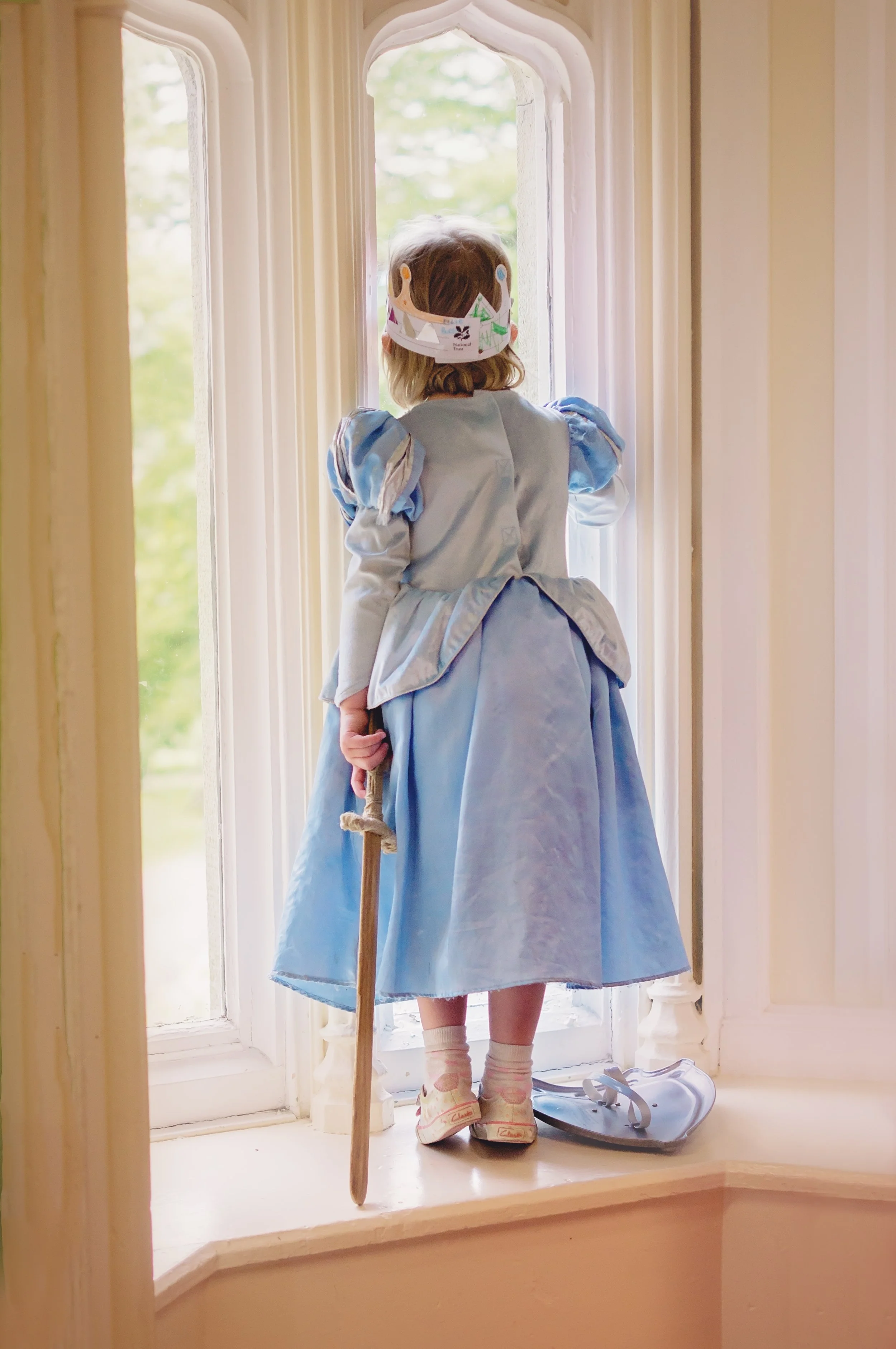 A young girl dressed as Cinderella standing on a window ledge, looking out the window. She wears a blue ball gown, a paper crown, and holds a small stick. A small blue bag is placed beside her on the ledge.