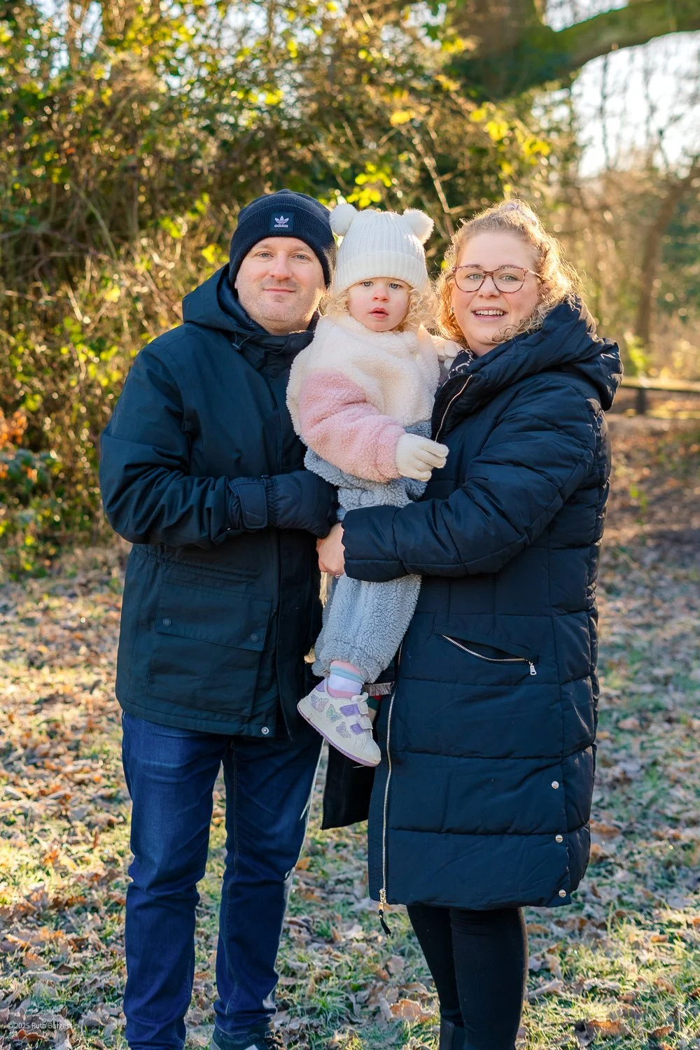 A family of three standing outdoors in a wooded area during fall, dressed warmly. The father and mother are holding their young daughter between them, all smiling at the camera.