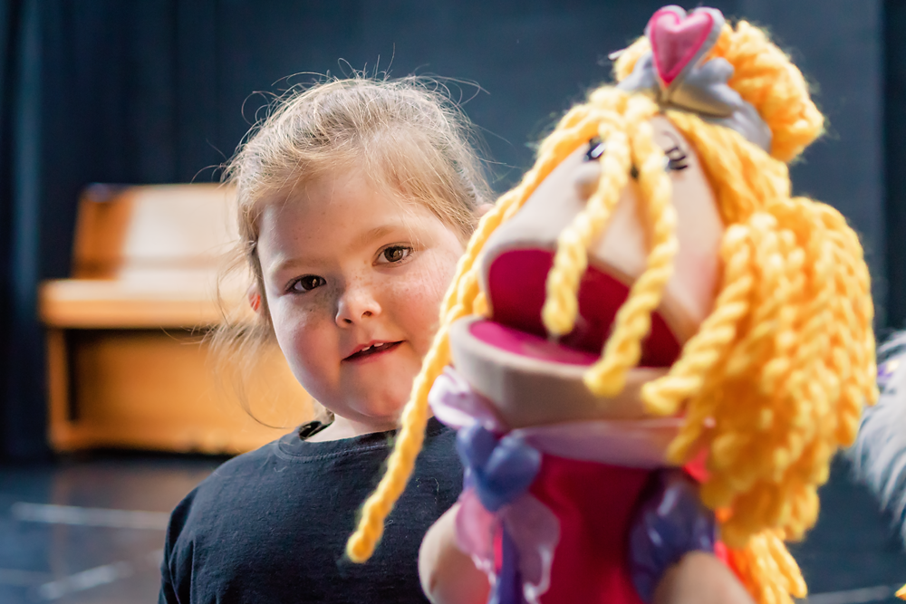Young girl holding a colorful sock puppet with yellow yarn hair and a pink bow, in a room with a piano in the background.