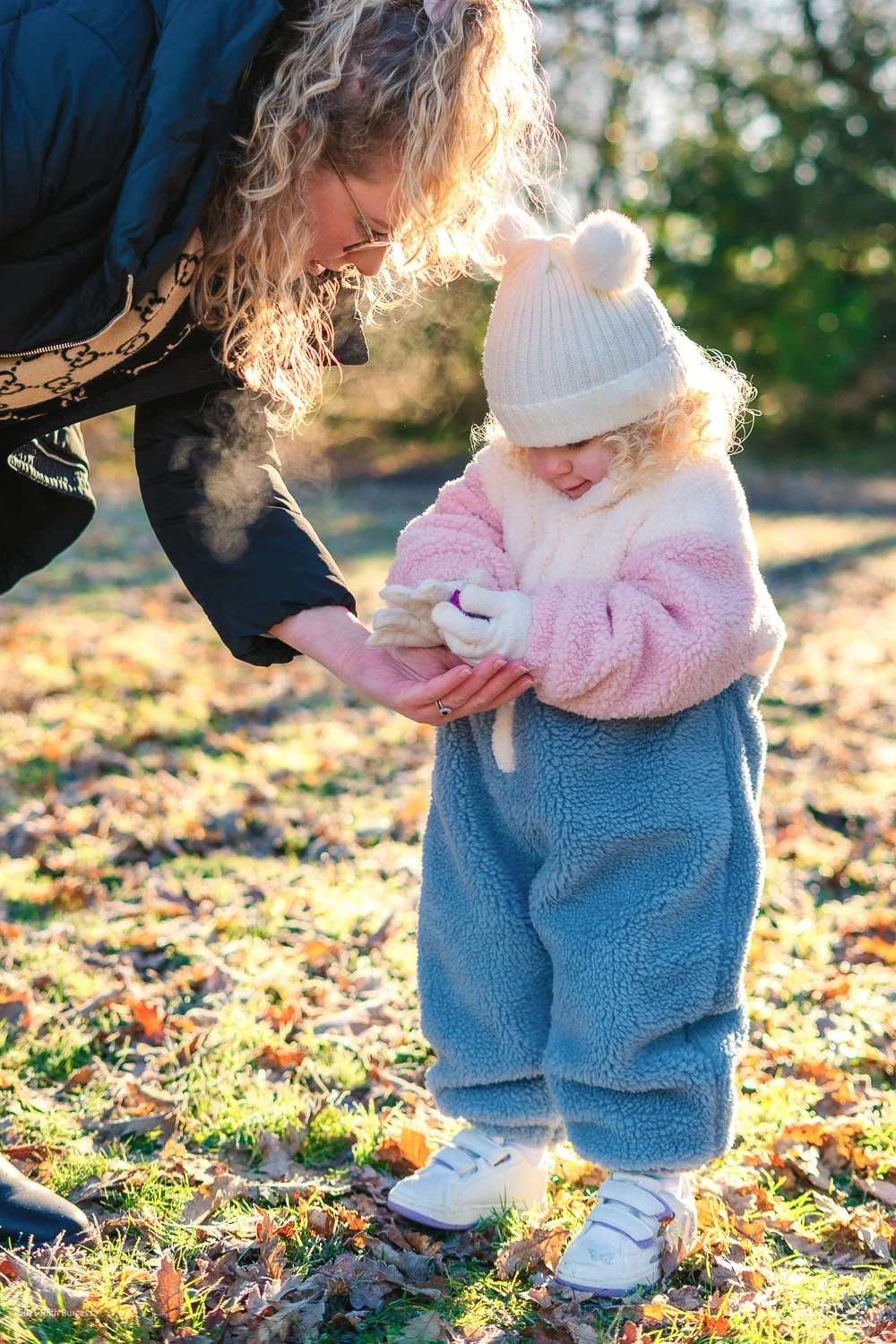 An adult woman and a young girl standing outdoors on a fall day, with the woman helping the girl put on mittens. Both are dressed warmly in fuzzy clothing and hats. The girl has a white knit hat with pom-poms and a pink and blue fuzzy outfit.
