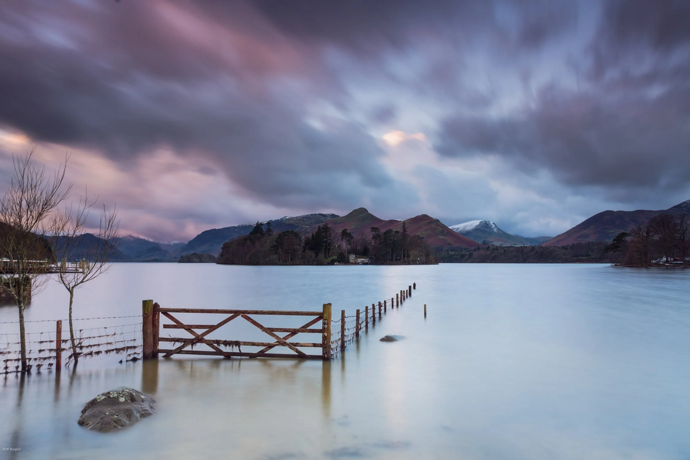 Scenic view of a lake with a wooden fence partially submerged in water, surrounded by hills and mountains with snow on some peaks, and a cloudy sky overhead during twilight.