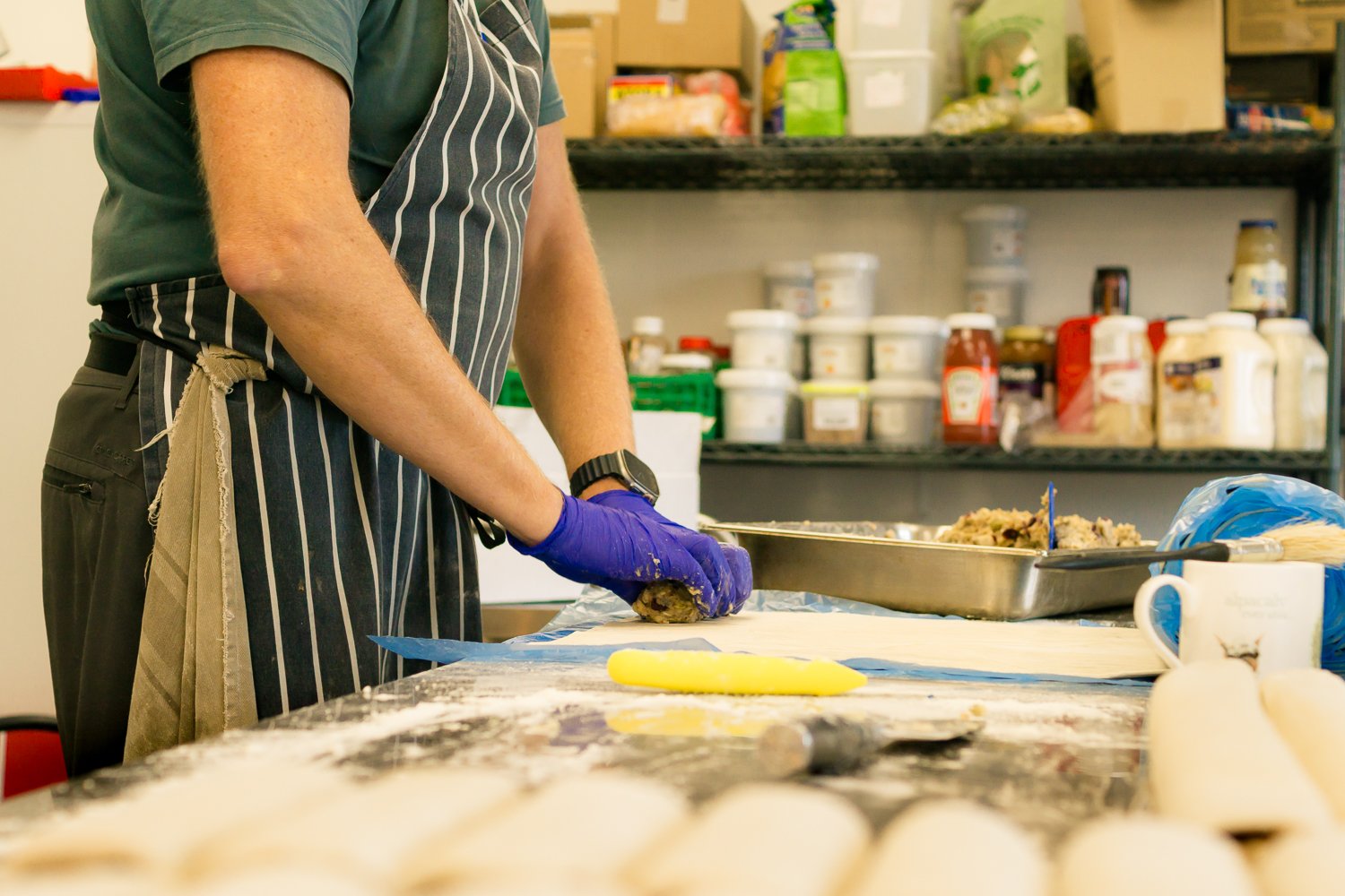 Person wearing a striped apron and purple gloves wrapping stuffed dough in a kitchen or bakery setting.