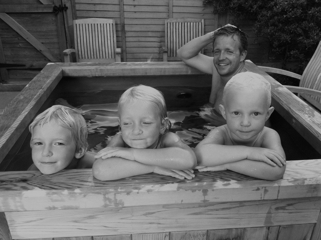 A man and three young children relaxing together in a wooden hot tub in a garden, smiling.
