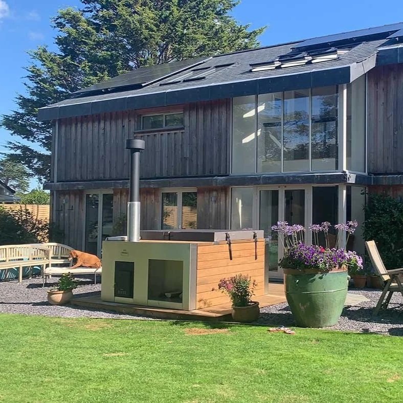 Backyard patio with outdoor dining table, chairs, umbrellas, potted plants, a Tubmarine Hot Tub and a wooden house with large windows. 