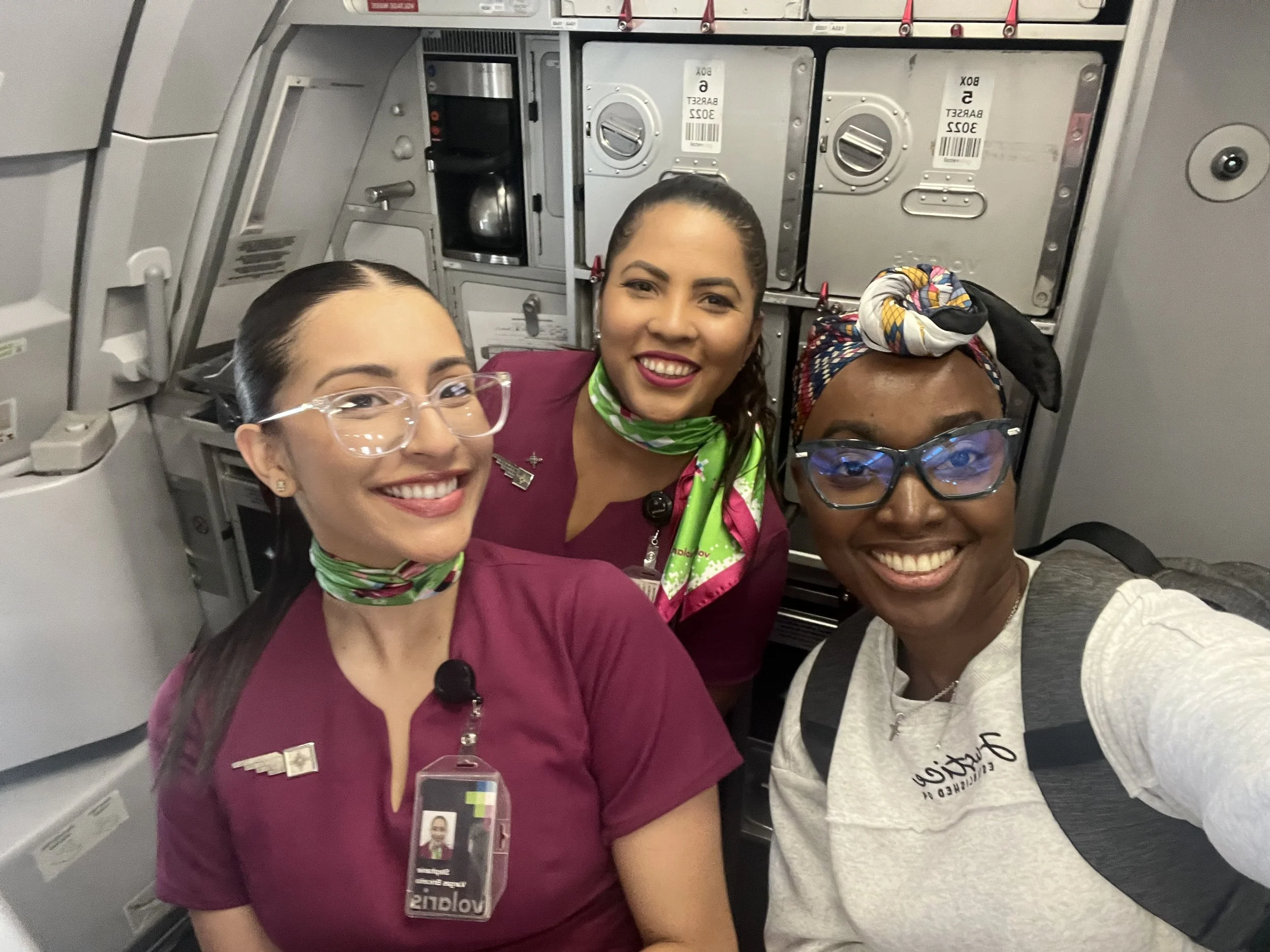 Three smiling women, two flight attendants and one passenger, taking a selfie inside an airplane cabin.