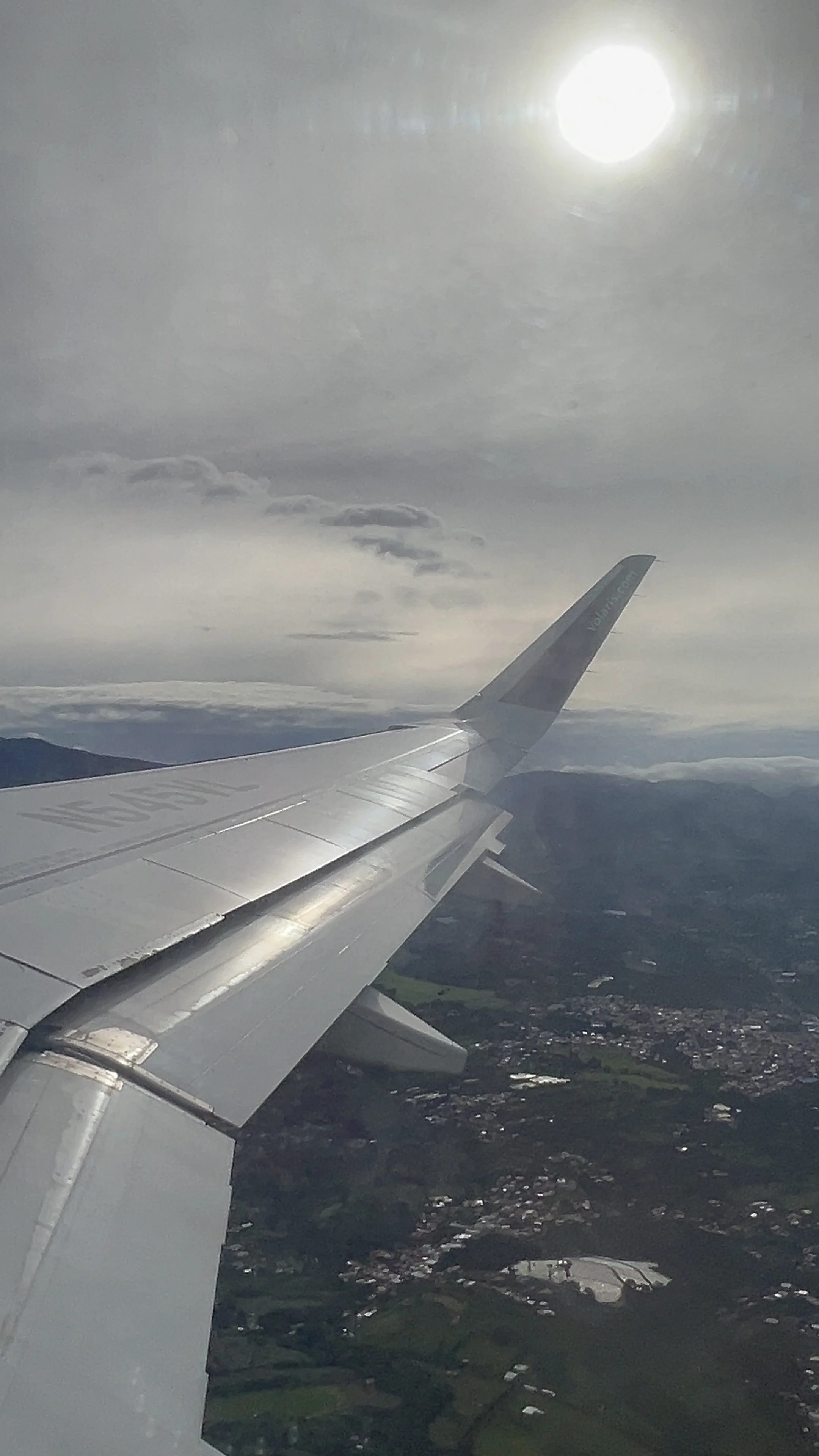 View from an airplane window showing the wing of the plane, clouds, the sun, and landscape below.