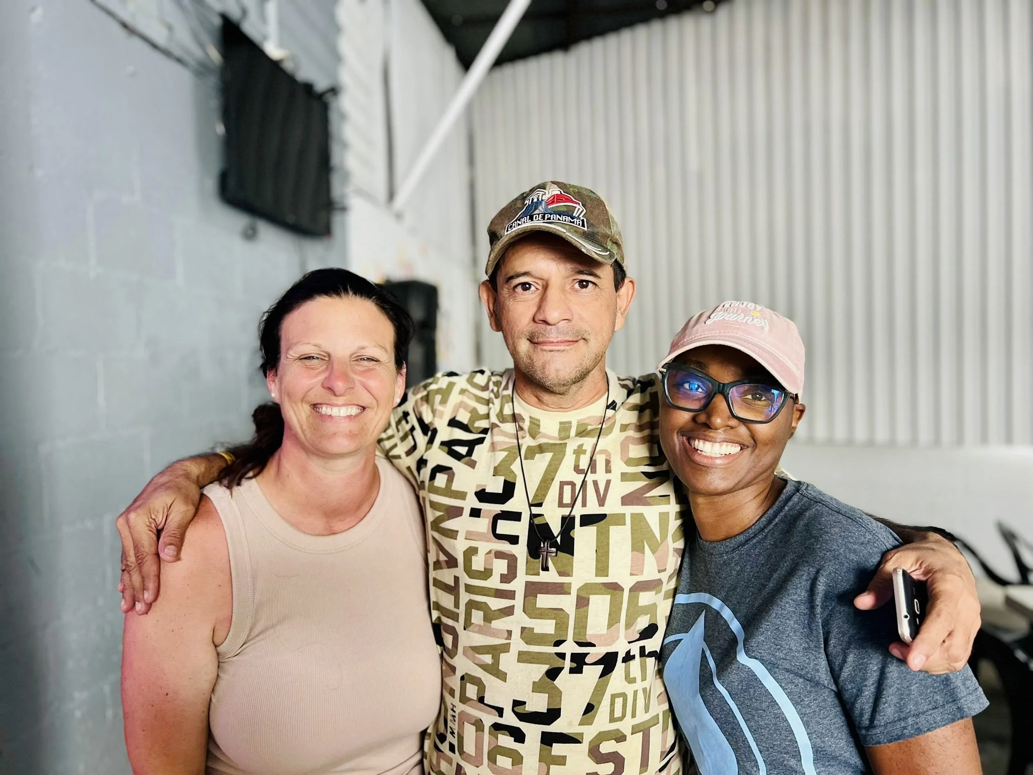 Three people standing close together with arms around each other, smiling at the camera in a room with gray walls and metal siding, two women and one man, casually dressed in T-shirts, with the woman on the right wearing glasses and a pink cap.