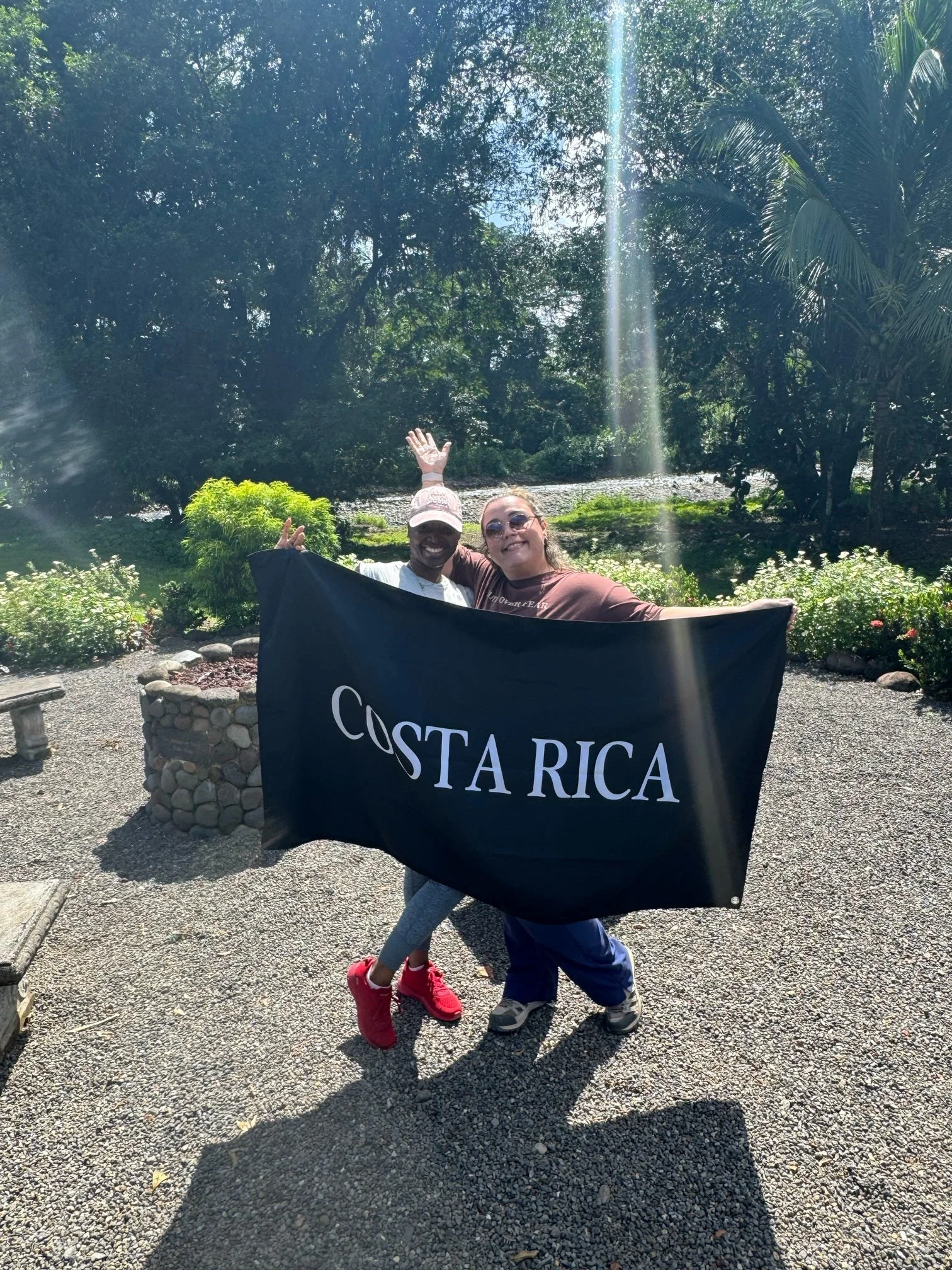 Two children are smiling and holding a large black flag with the words "COSTA RICA" on it, outdoors in a lush park with trees, sunlight, and a small stone border around the flowerbed.