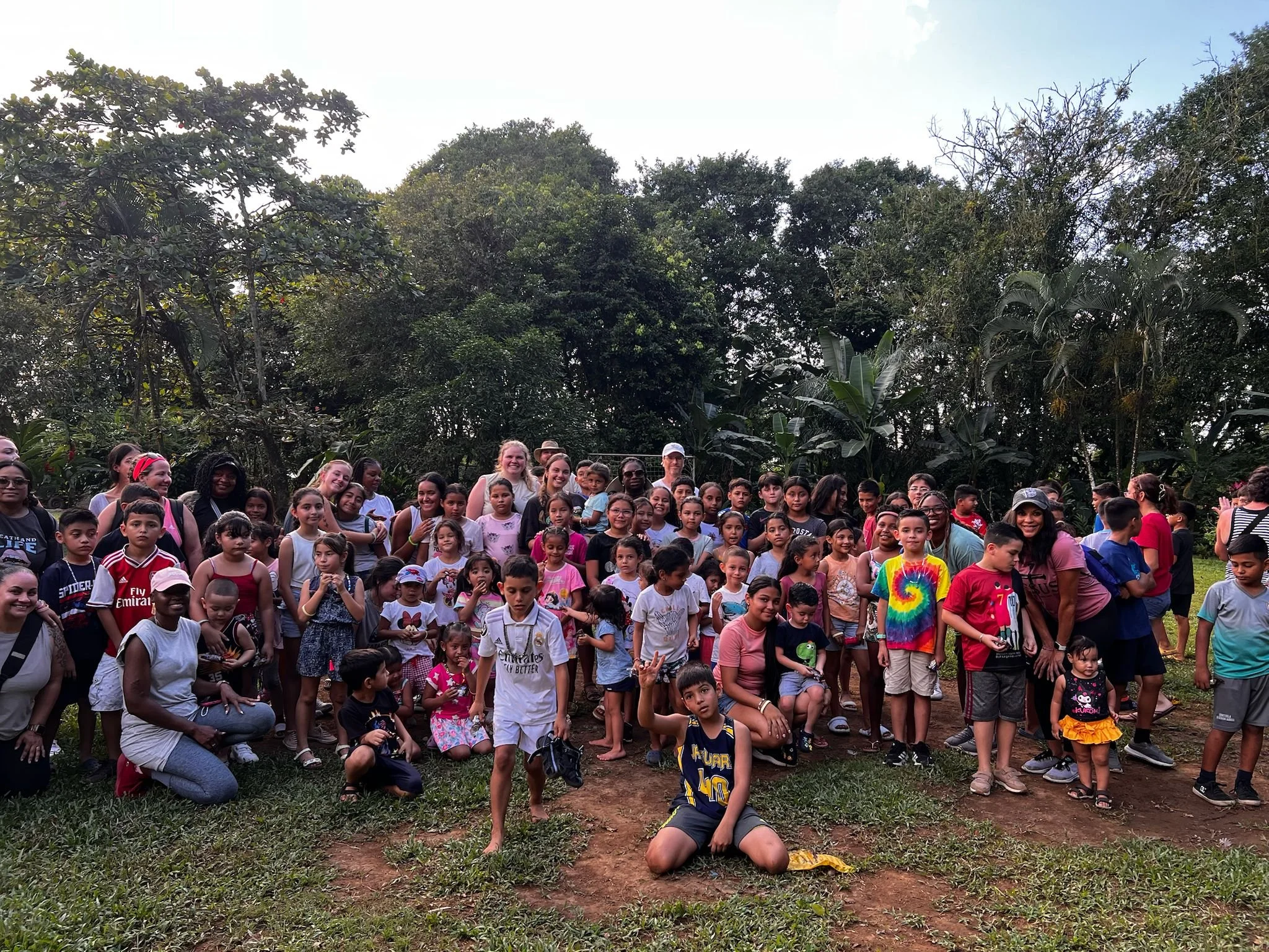 Large group of children and adults gathered outdoors in a lush green area with trees in the background.