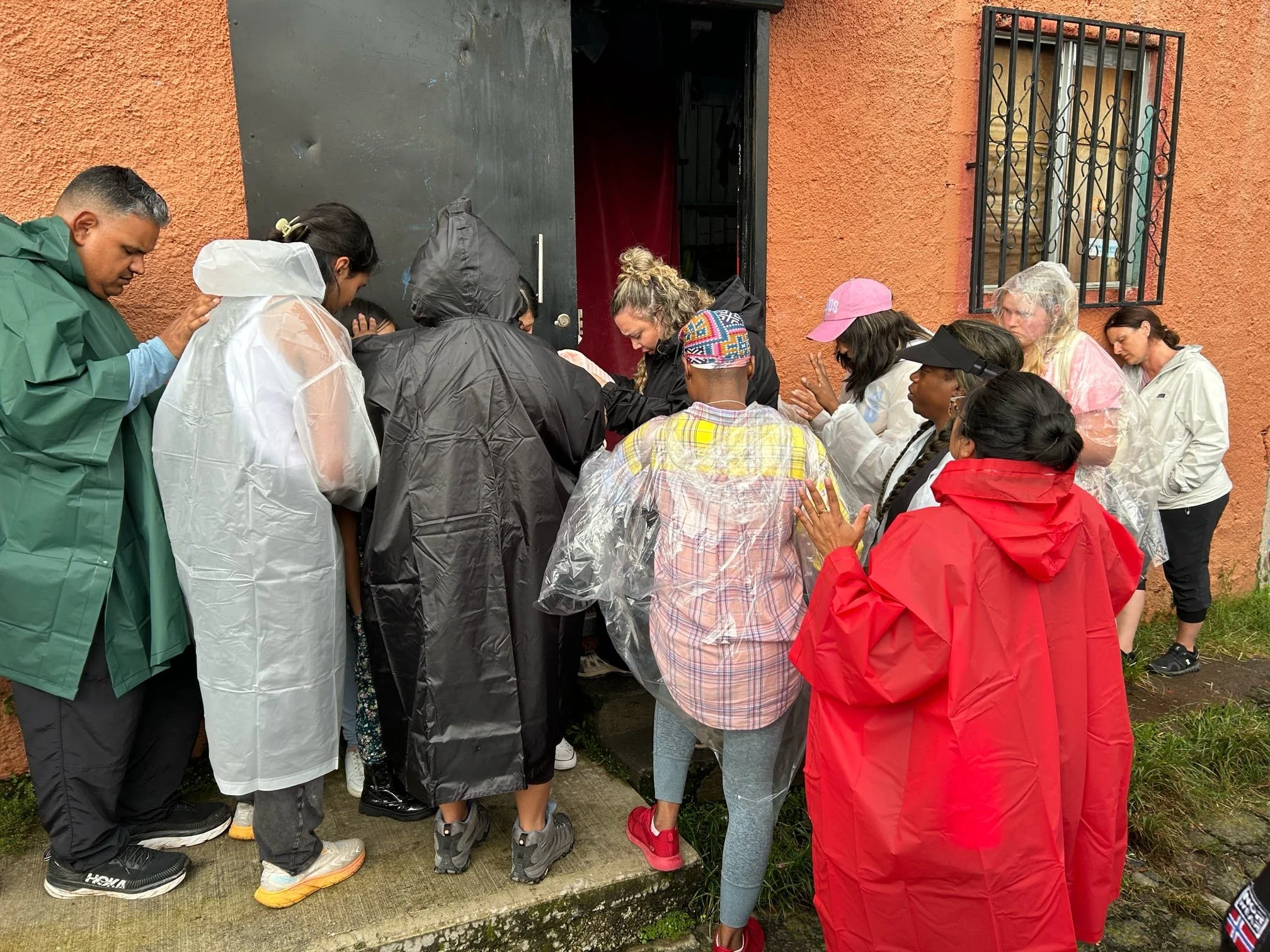 Group of people gathered outside a building with orange walls, some wearing rain ponchos, as they participate in a religious or spiritual activity near a doorway.