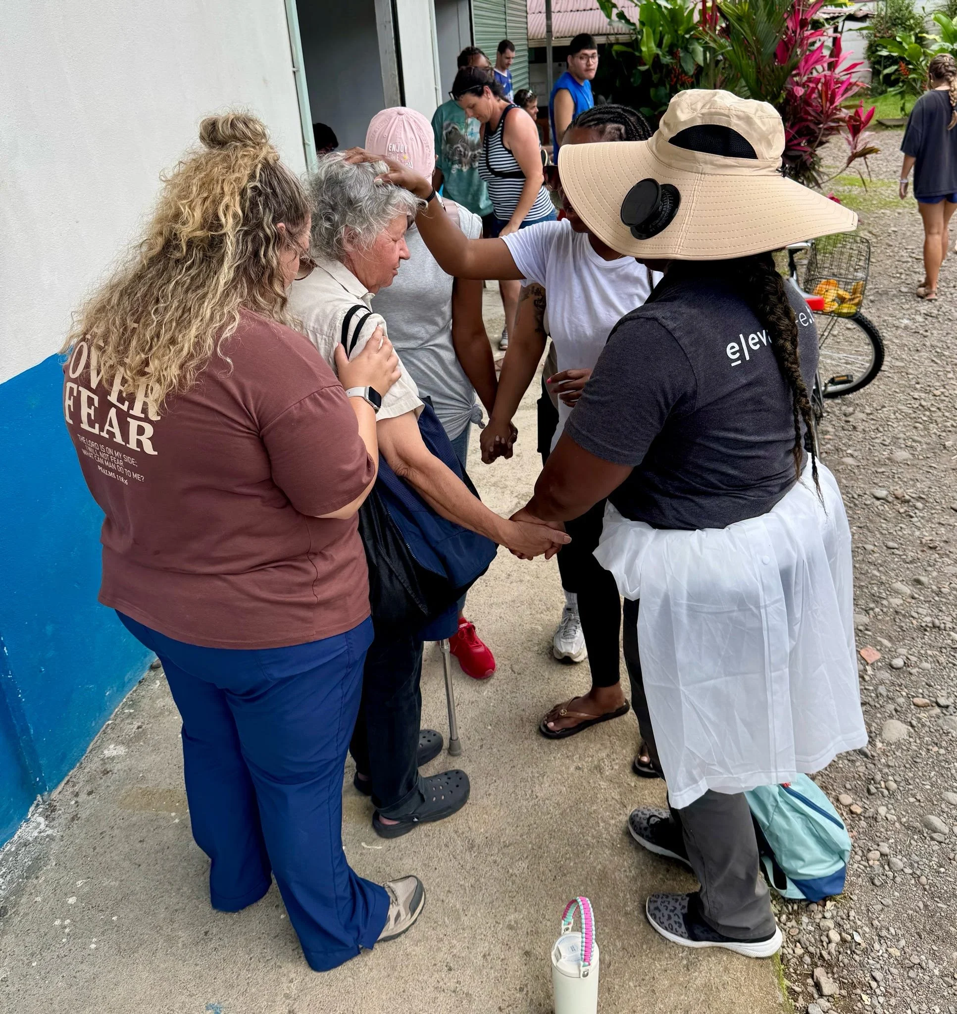 Group of people praying outside, holding hands, with some people wearing hats and backpacks, near a building with plants in the background.