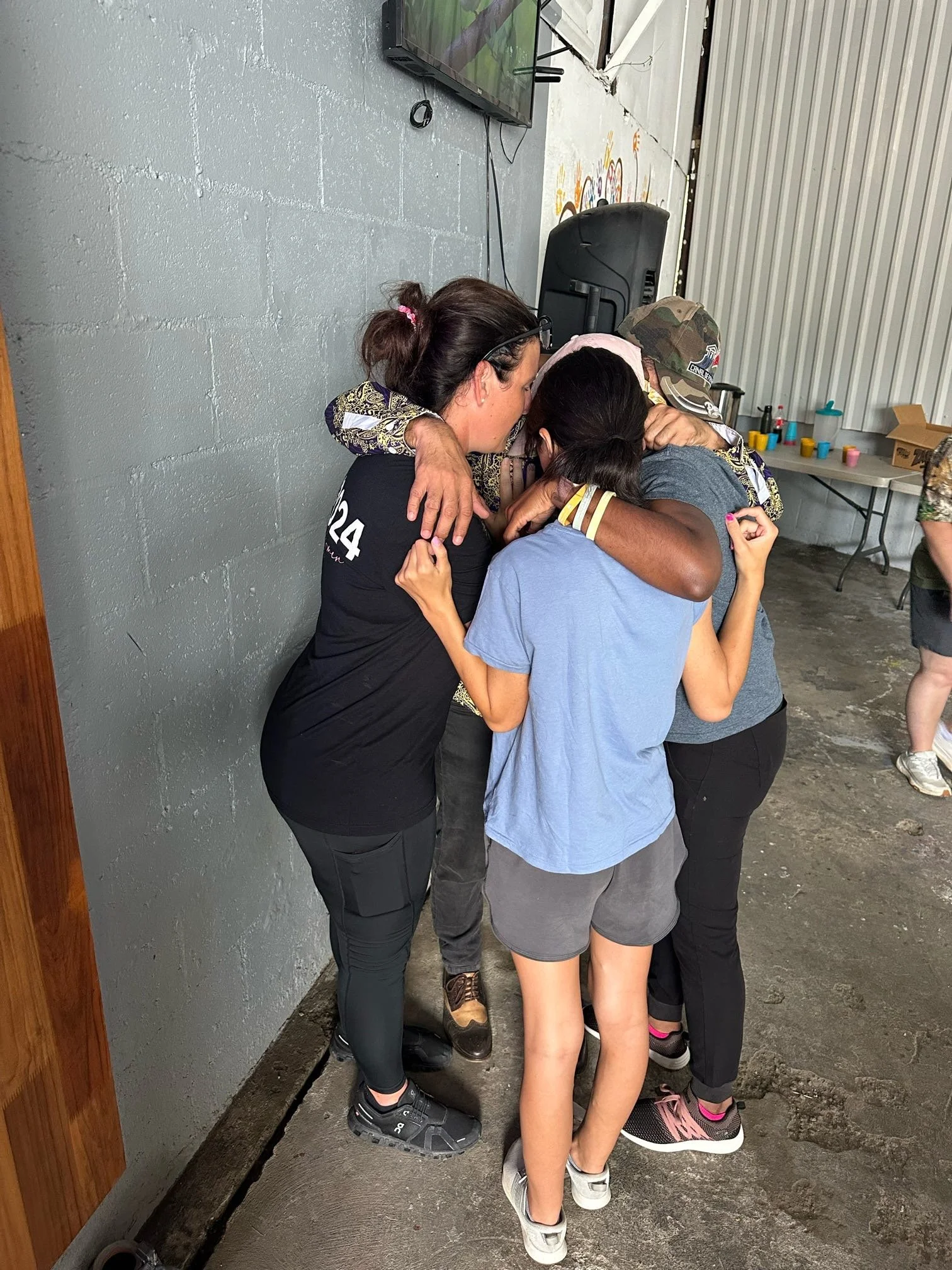 Group of four people in a huddle, embracing in an indoor setting with a gray wall and a television mounted above.
