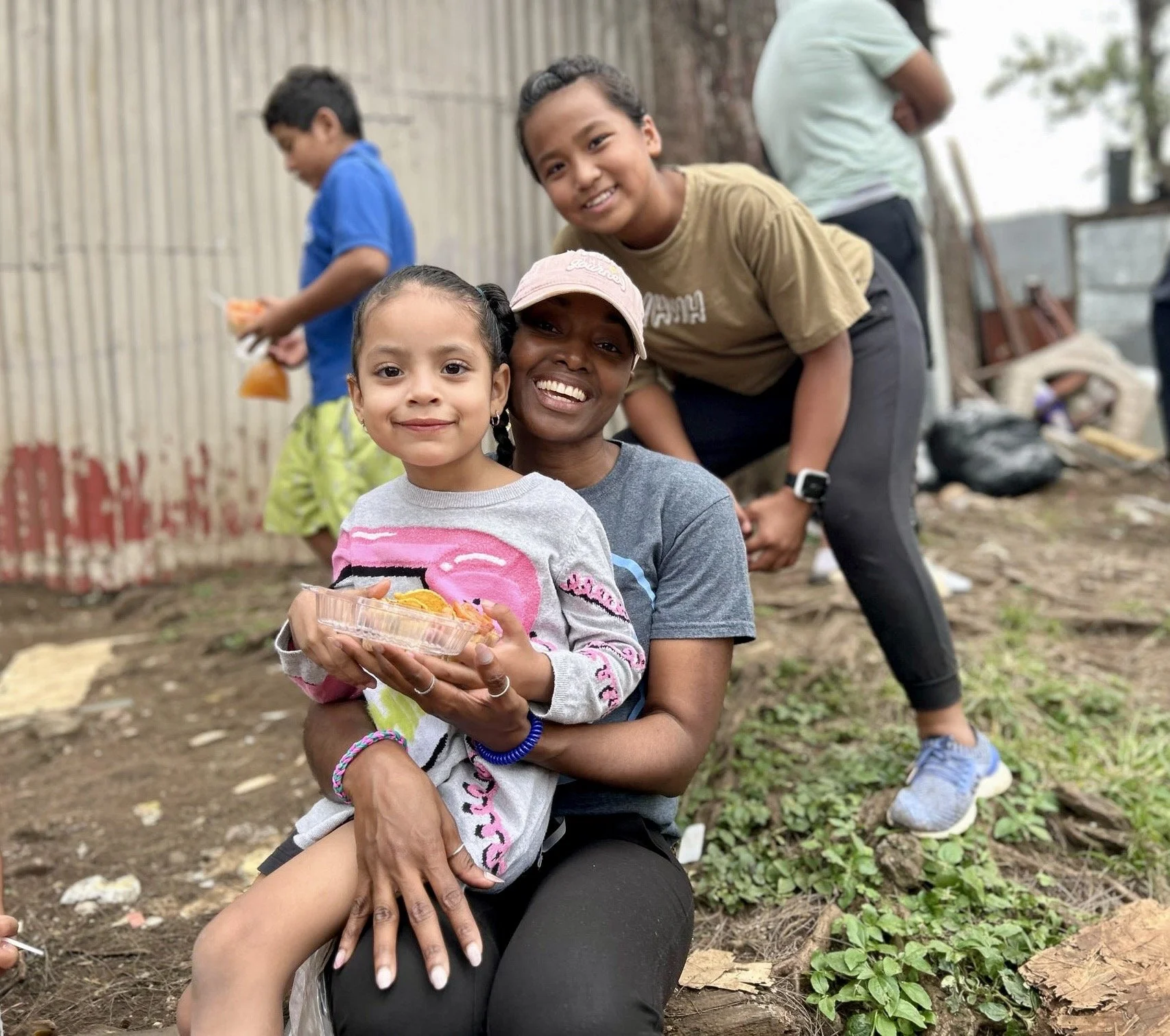A group of smiling people outdoors, including a woman holding a young girl with a container of food, others in the background, with a dirt ground and a wooden fence.