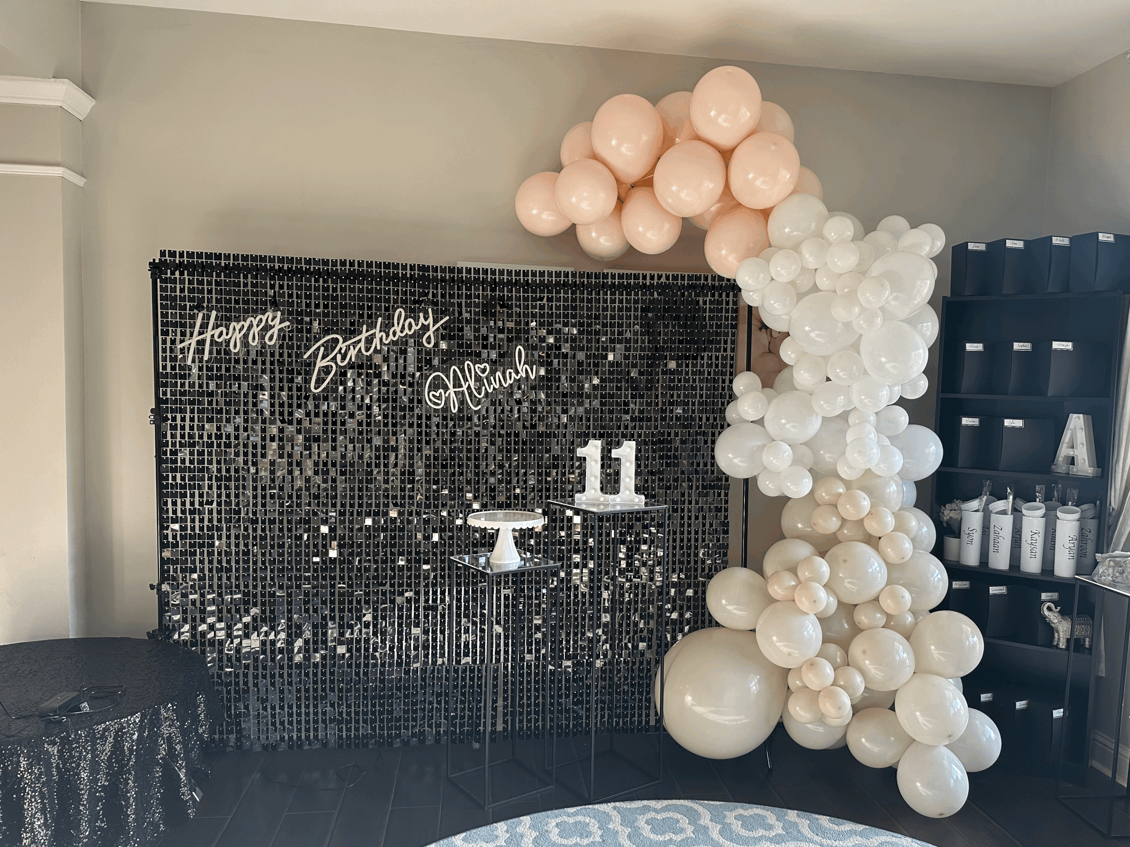 Decorative black mosaic backdrop with neon signs that read 'Happy Birthday Alinah.' A balloon arrangement with white and blush pink balloons cascading from the top right corner. A small table with a white cake stand holding a white number 11. A black shelving unit with labeled white cups and decorative items on the right side.
