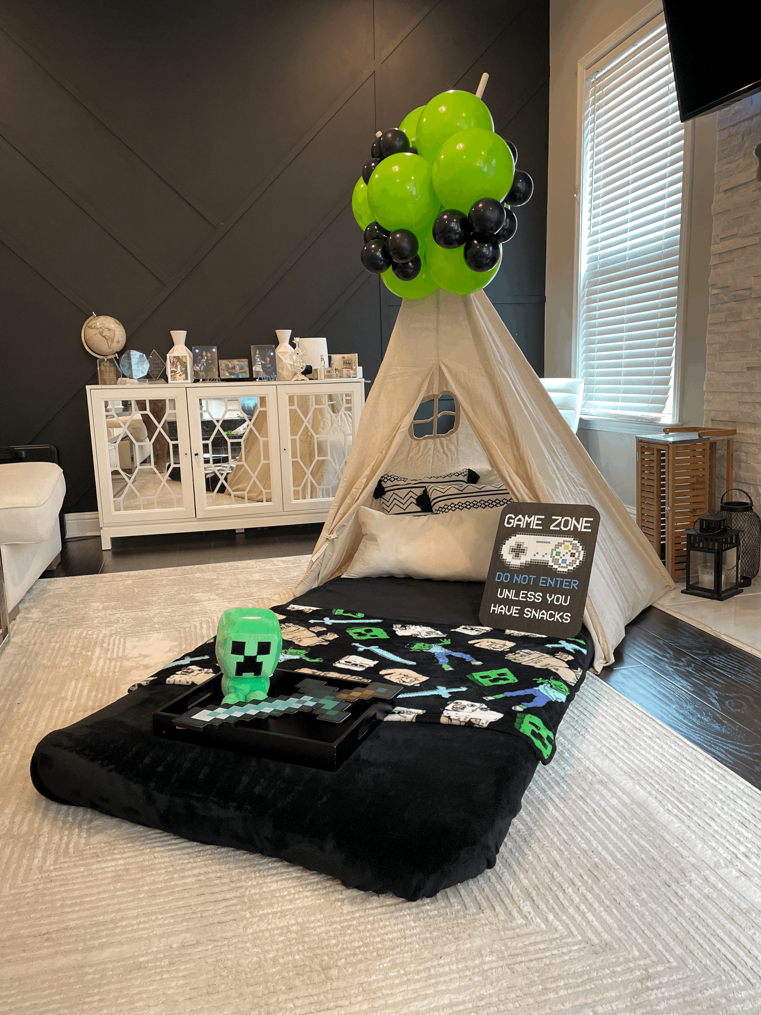 Indoor play area featuring a small teepee tent decorated with green and black balloons, a sign reading 'Game Zone Do Not Enter Unless You Have Snacks,' and Minecraft-themed decorations on a mattress with plush toys and pixelated figures. A tray with Minecraft figures and a Creeper lamp are in the foreground.