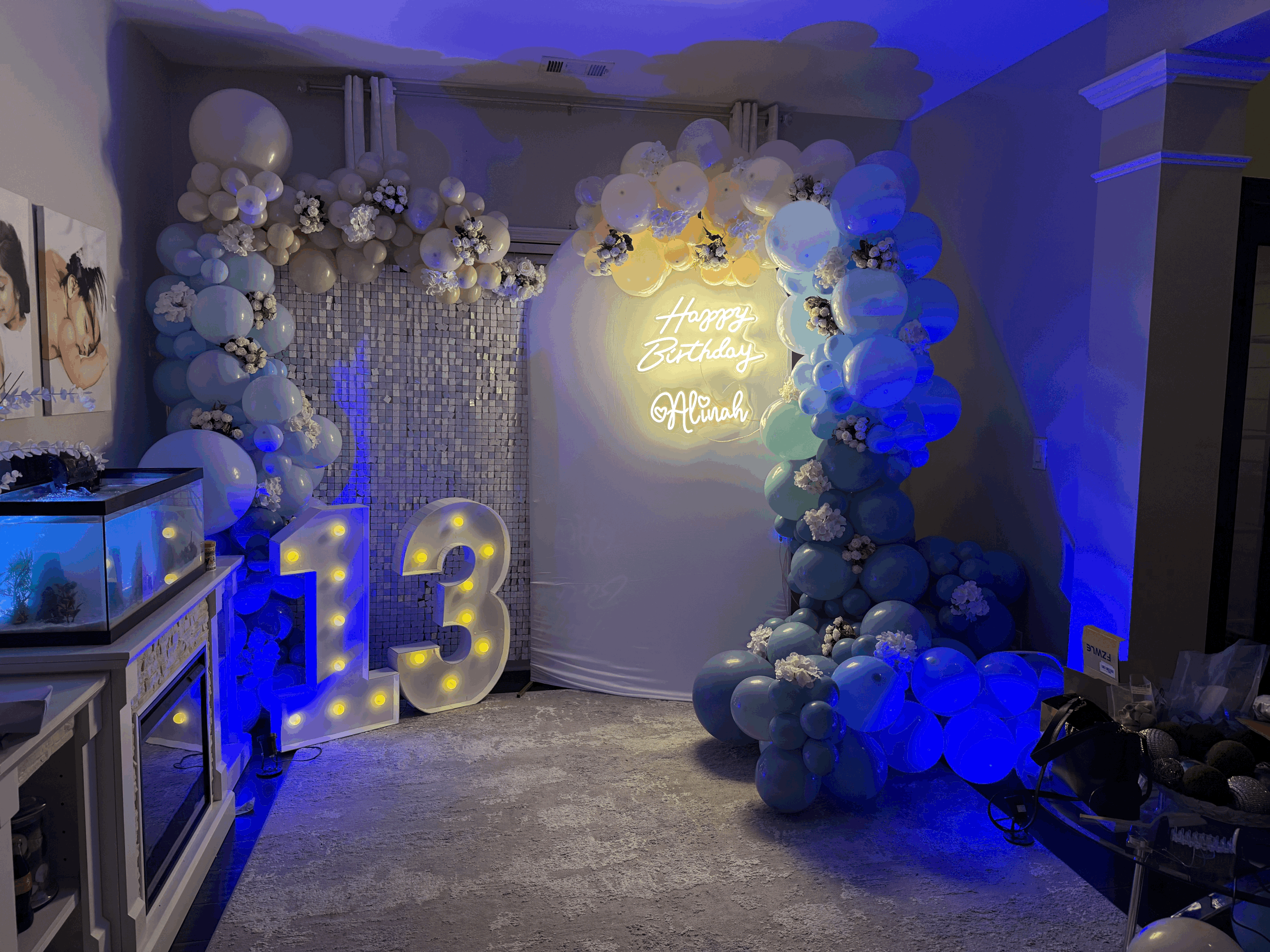 Decorated birthday celebration space featuring a large helium balloon arch, illuminated signs with the words 'Happy Birthday Alinah,' large lit-up number 13, and various smaller balloons, with a white textured backdrop and photographs on the wall.