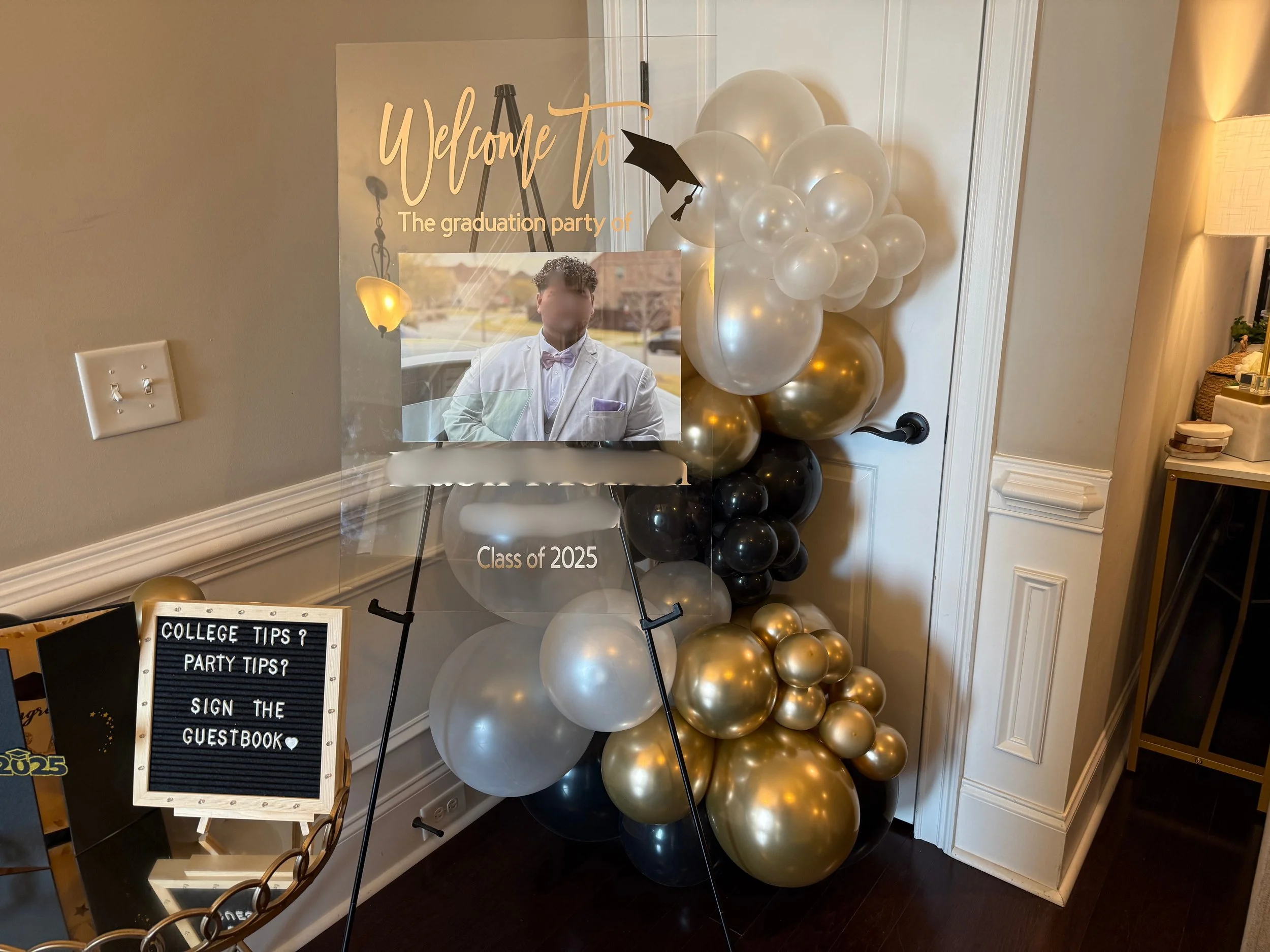 Decorative setup for a graduation party, featuring a wall sign with a photo of a young man in a white tuxedo, a cluster of black, gold, and white balloons, a signboard with party tips, and a transparent acrylic stand with the words 'Welcome to the graduation party of Class of 2025'.