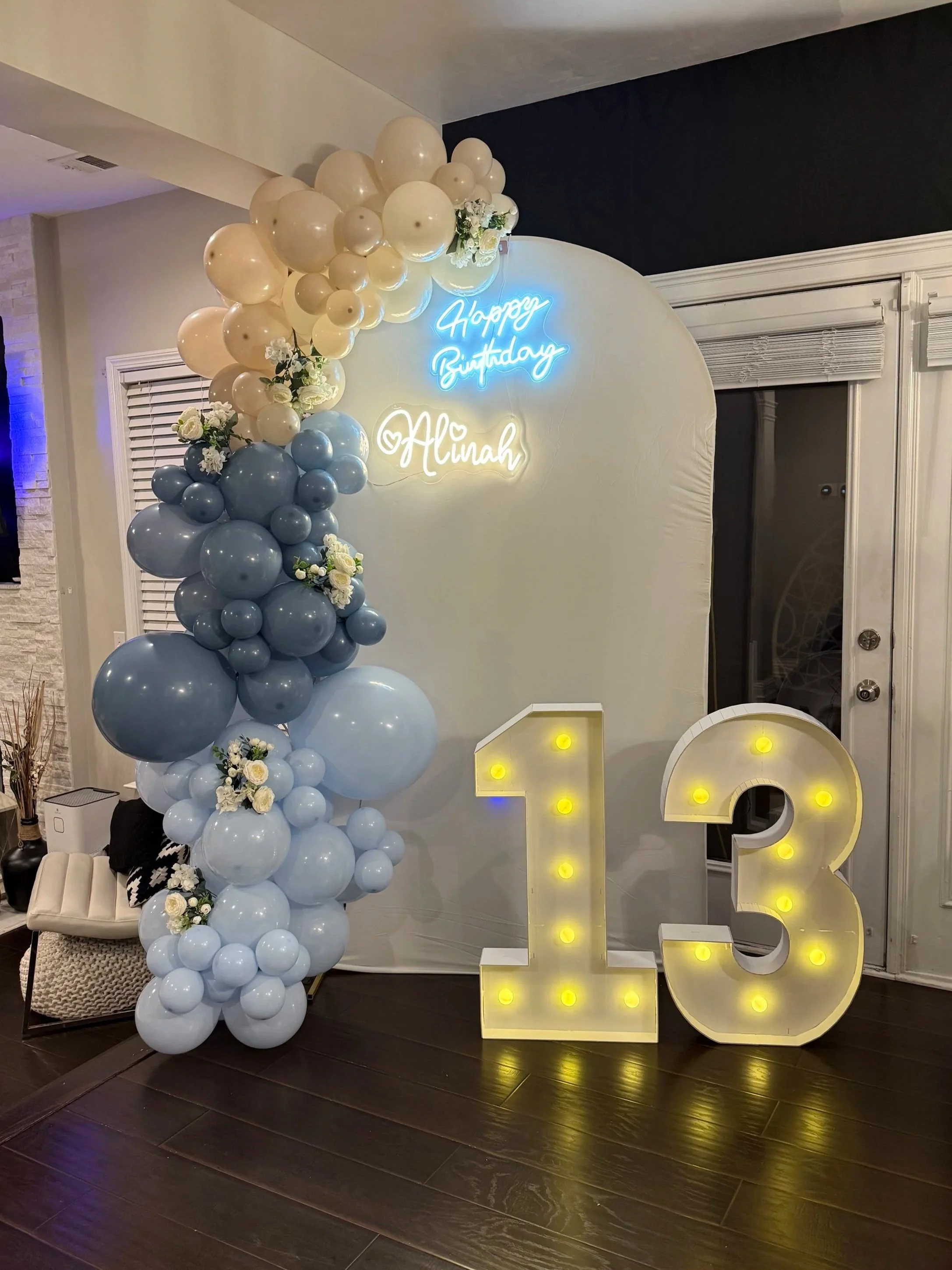 Decorative birthday celebration setup with a white backdrop, a vertical arrangement of cream and blue balloons with flowers, neon signs saying 'Happy Birthday Alinah', and large illuminated number '13' on the floor.