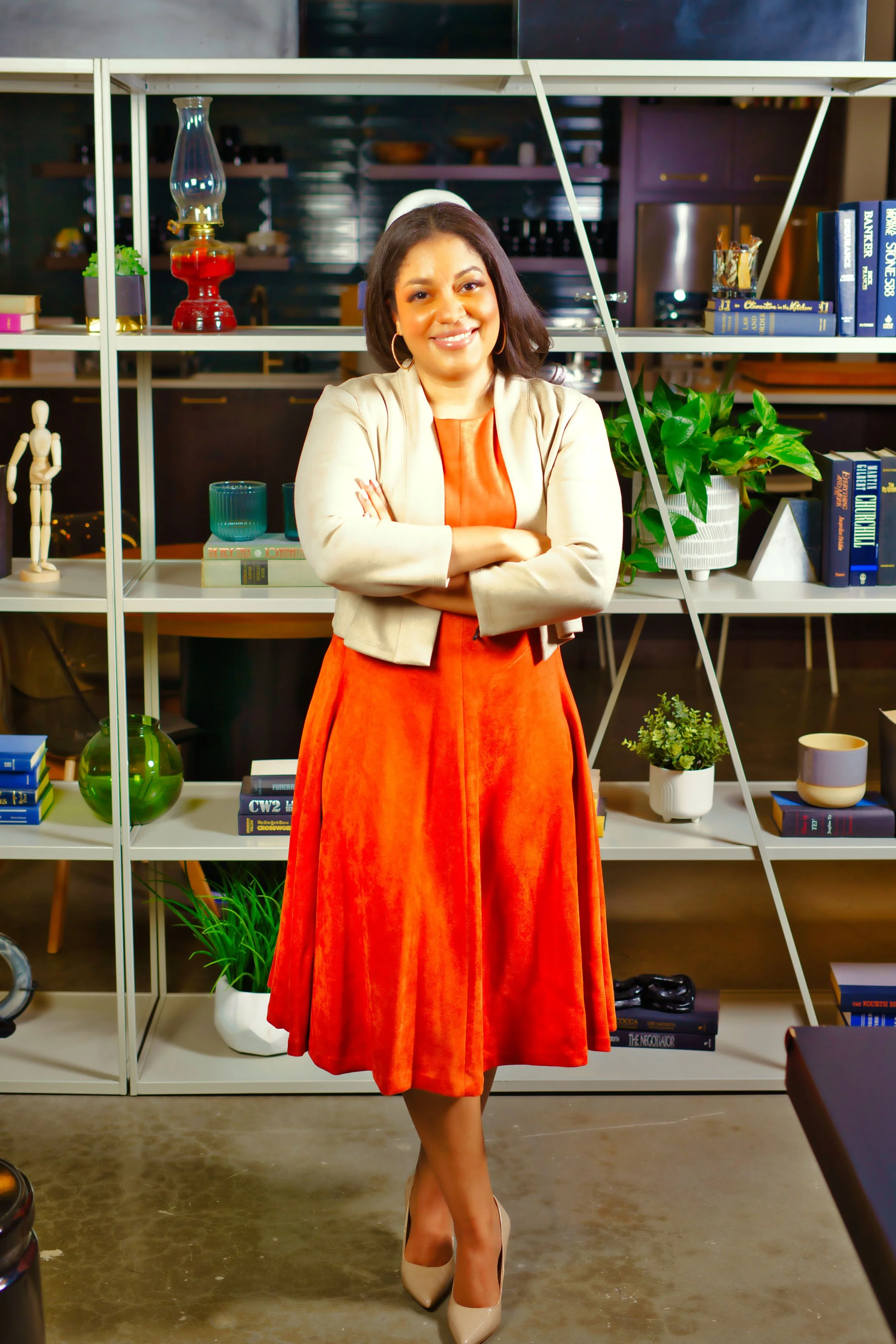 A woman standing with arms crossed in front of a bookshelf in a cozy, well-decorated room.