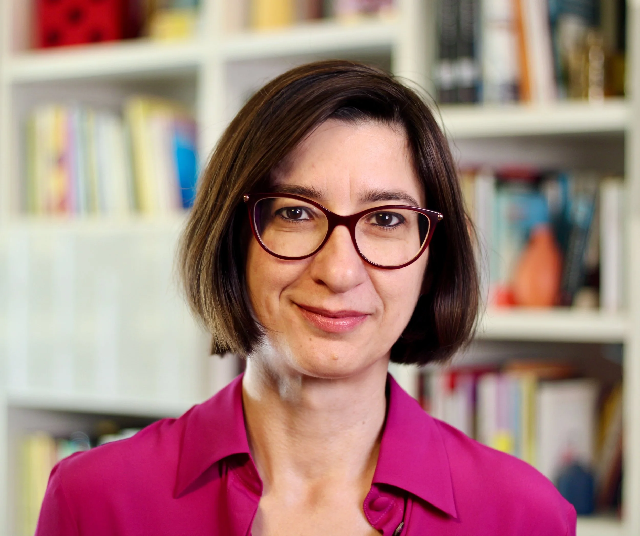 Donna con capelli castani, occhiali rossi e maglietta rosa davanti a una libreria.