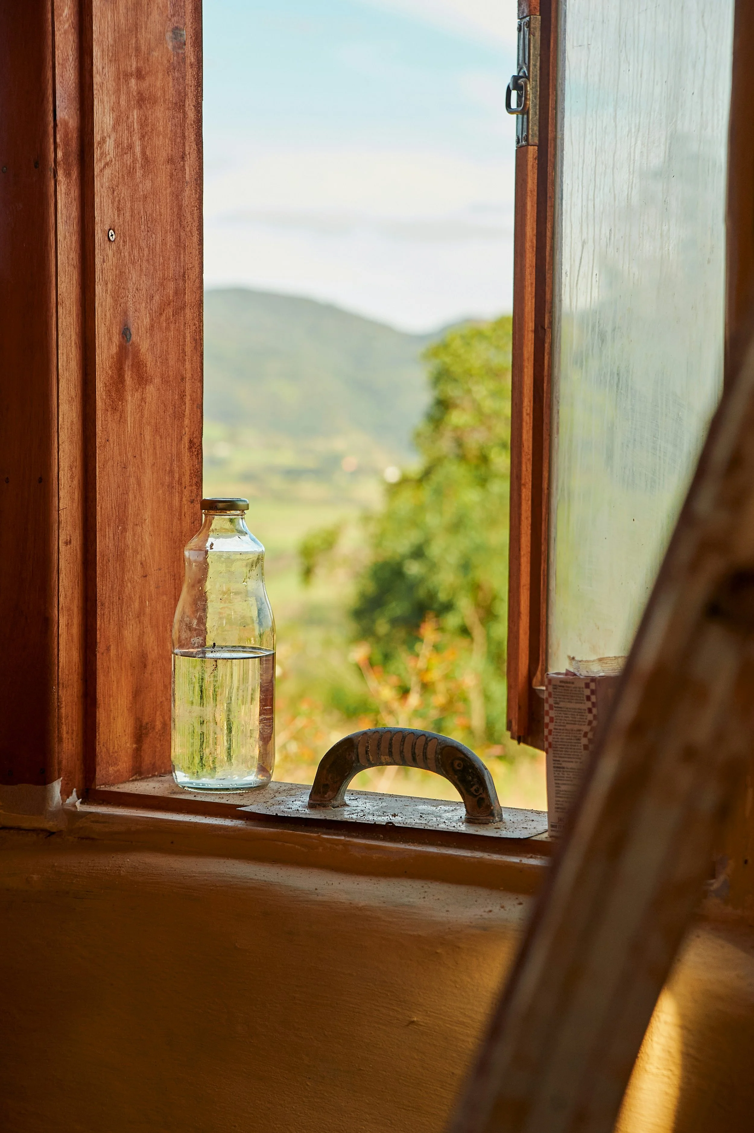 Finestra di legno aperta con vista sul paesaggio naturale e collina, bottiglia di vetro con acqua sulla finestra, oggetti da cucina e parte di una scala in primo piano.