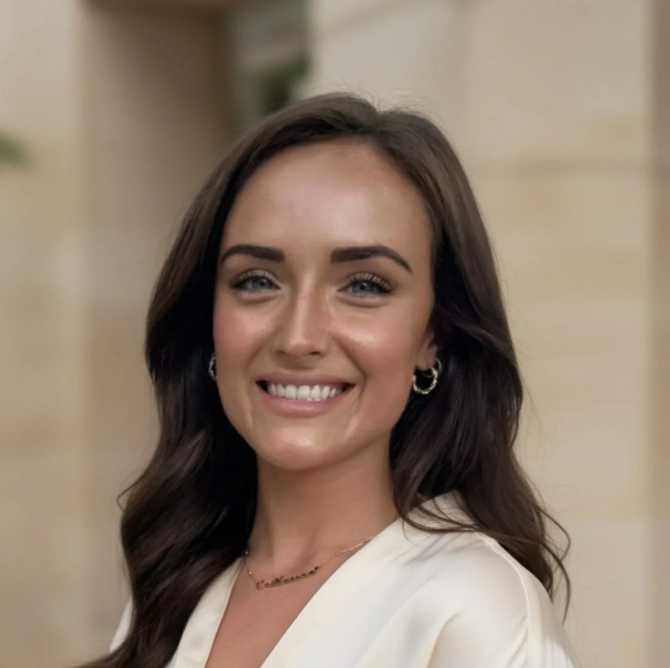 Close-up of a smiling woman with long dark hair, blue eyes, wearing gold hoop earrings and a white top, in an indoor setting.