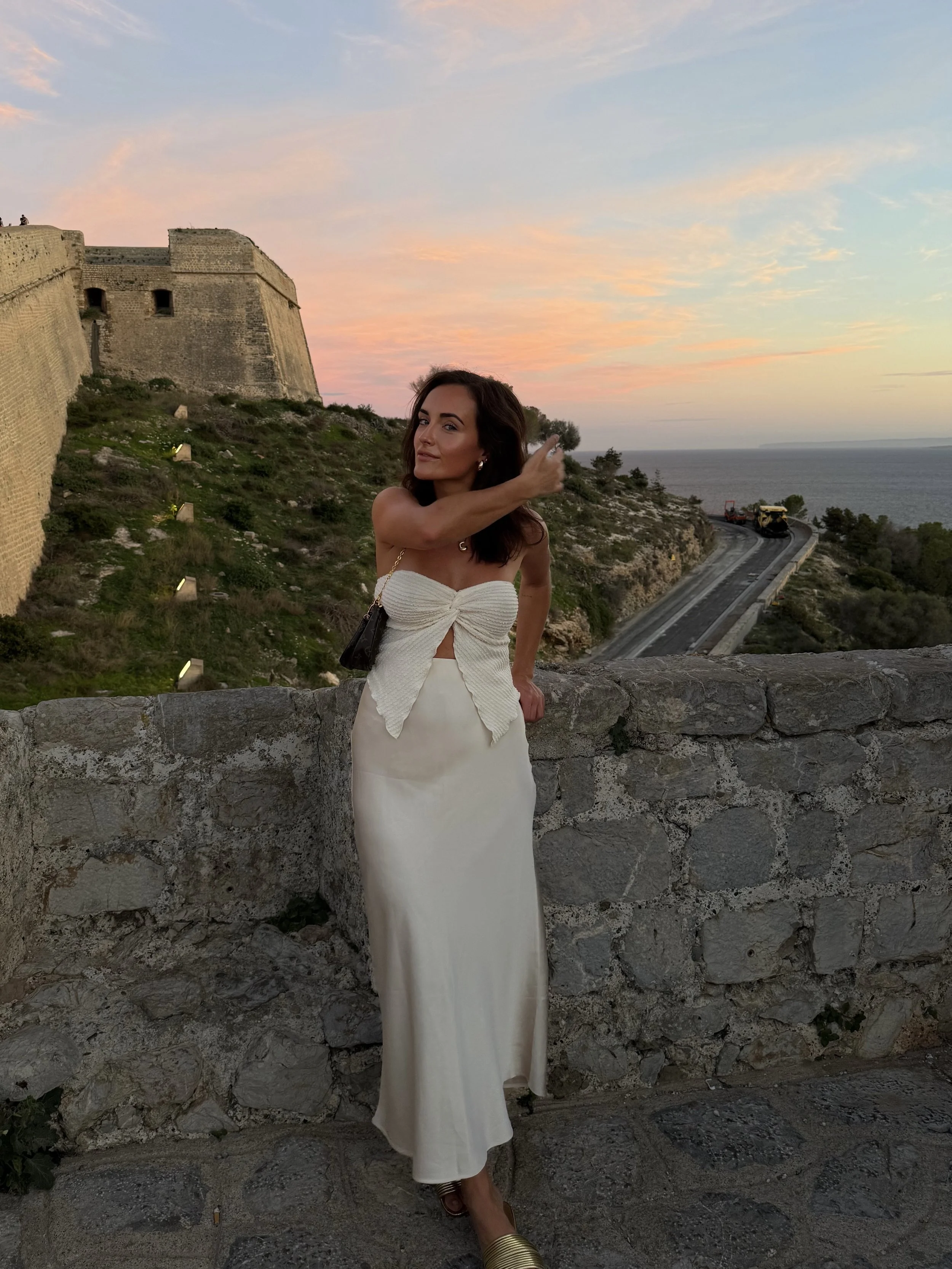 A woman in a white dress standing against a stone wall at sunset with a fortress and the sea in the background. Ibiza old town castle.