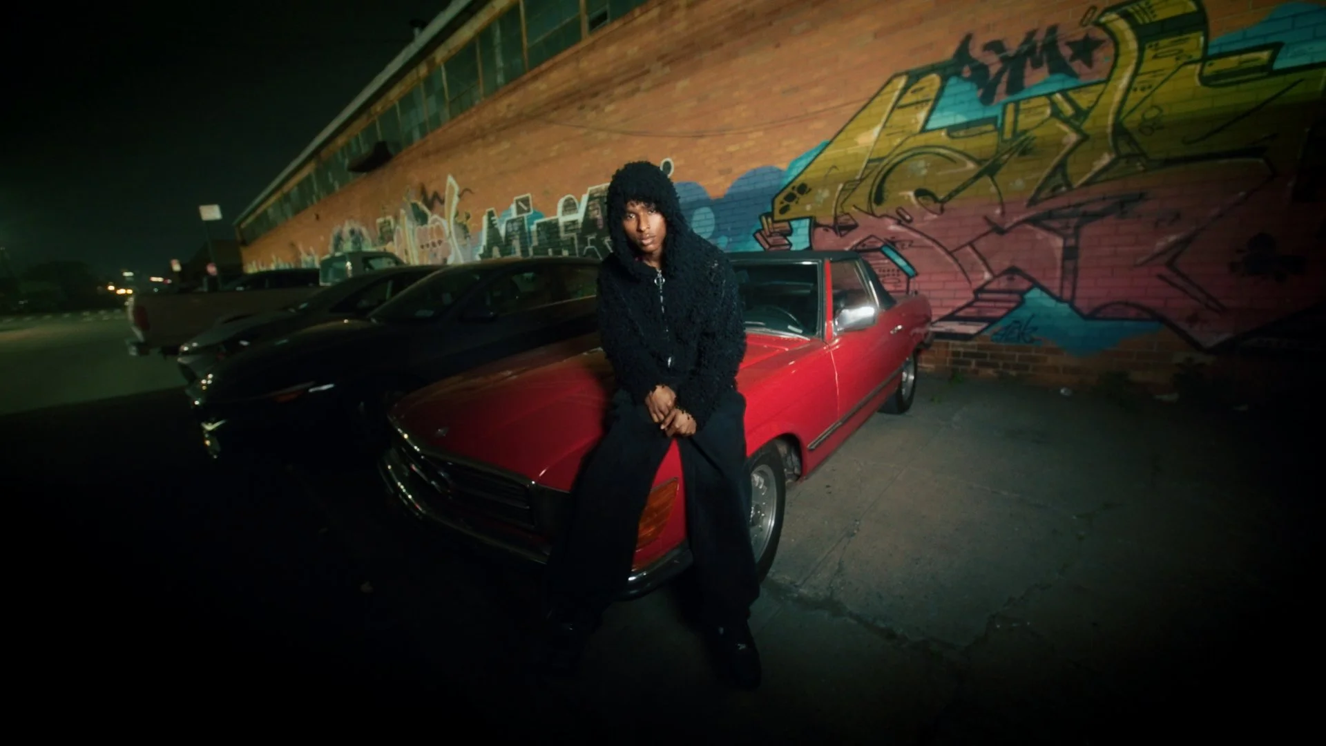 A person wearing a black fuzzy hoodie and black pants is sitting on the front of a red vintage car, parked in a lot at night. Behind them is a brick wall with colorful graffiti art.