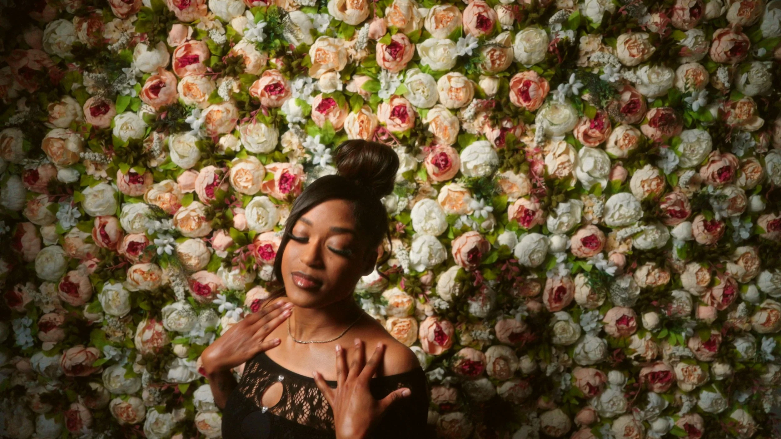 A woman with medium skin tone and dark hair styled in a bun stands in front of a wall decorated with pink and white roses and green leaves. She has her eyes closed and is touching her neck and shoulder area with both hands, wearing a black lace top with shoulder cutouts and a silver necklace.