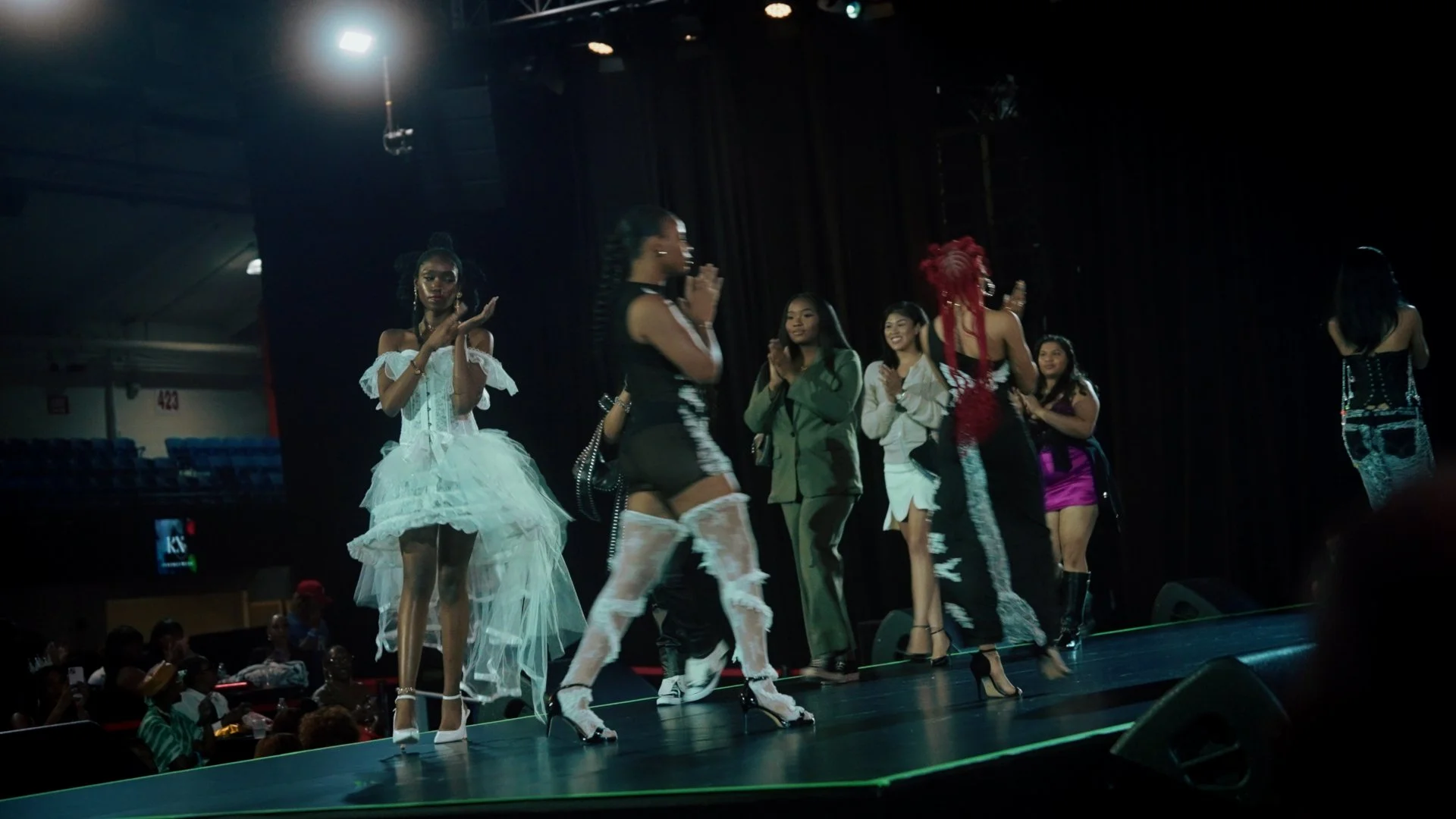 Group of women on stage at a fashion show, wearing various stylish outfits, some clapping, in a darkened venue with stage lighting.