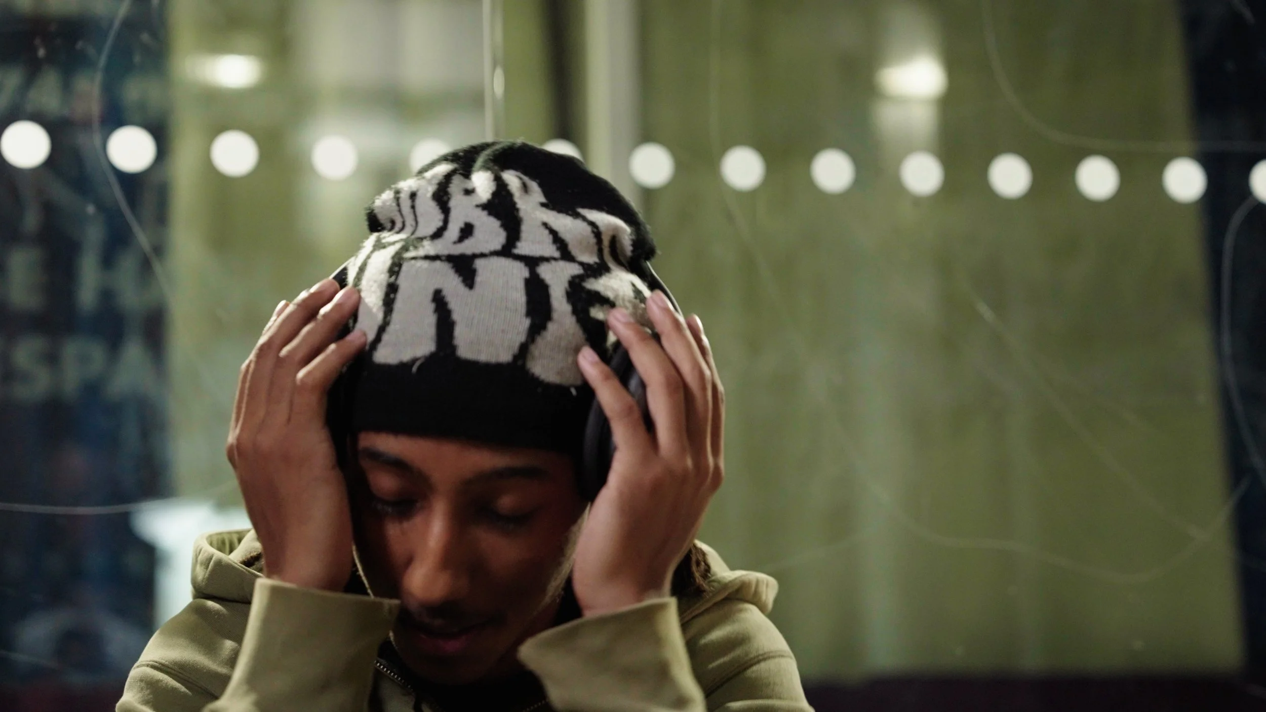 A person with dreadlocks and a patterned beanie, wearing headphones, holding their head with both hands, with eyes closed.