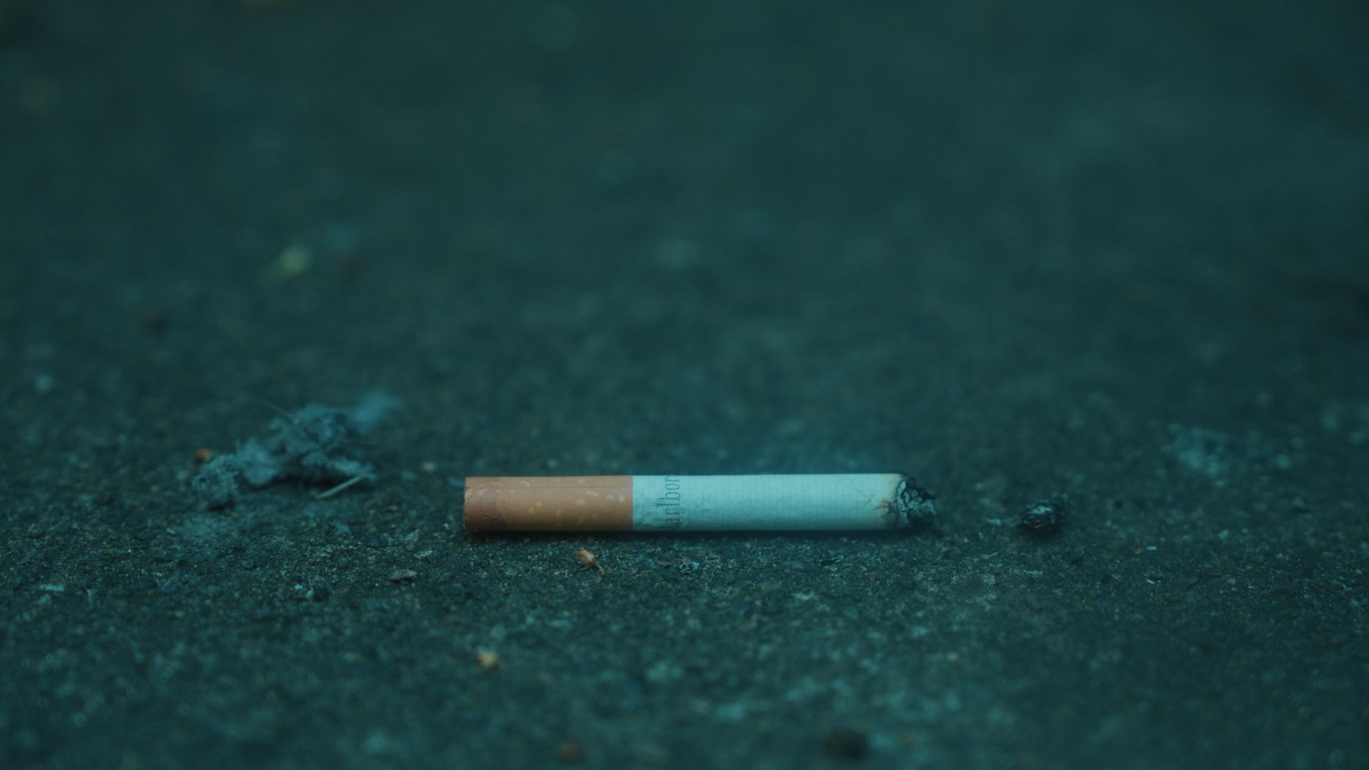 A discarded cigarette lies on the dark pavement, with ashes and debris nearby.