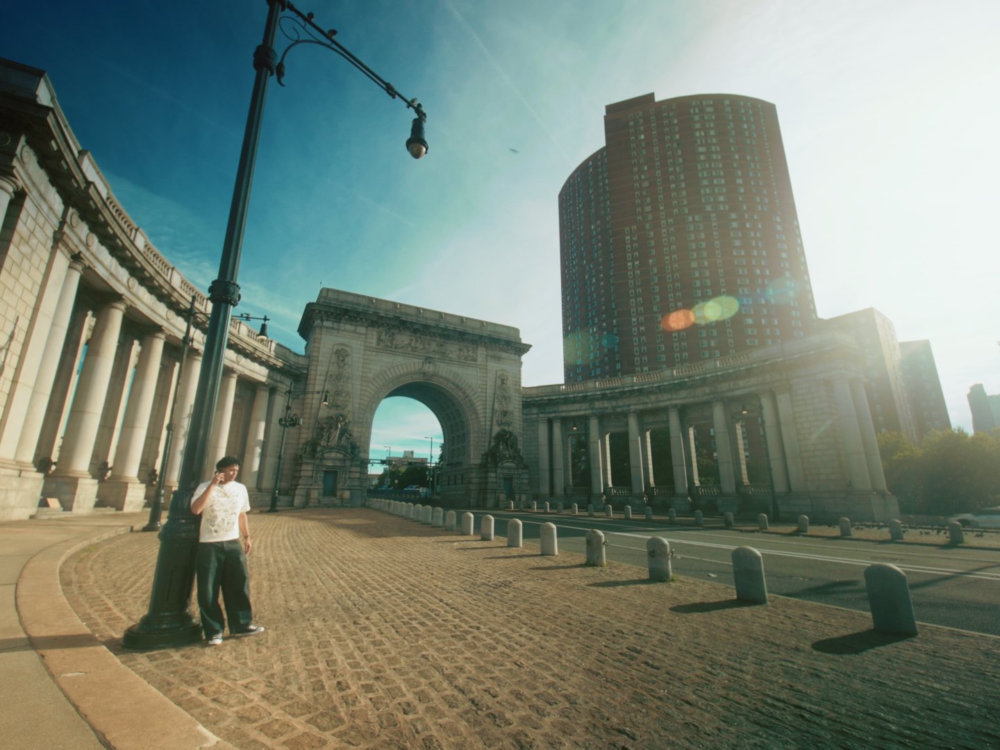 A woman leaning against a streetlamp in front of an arched monument and classical columns with a tall modern building in the background, sunny sky with lens flare.