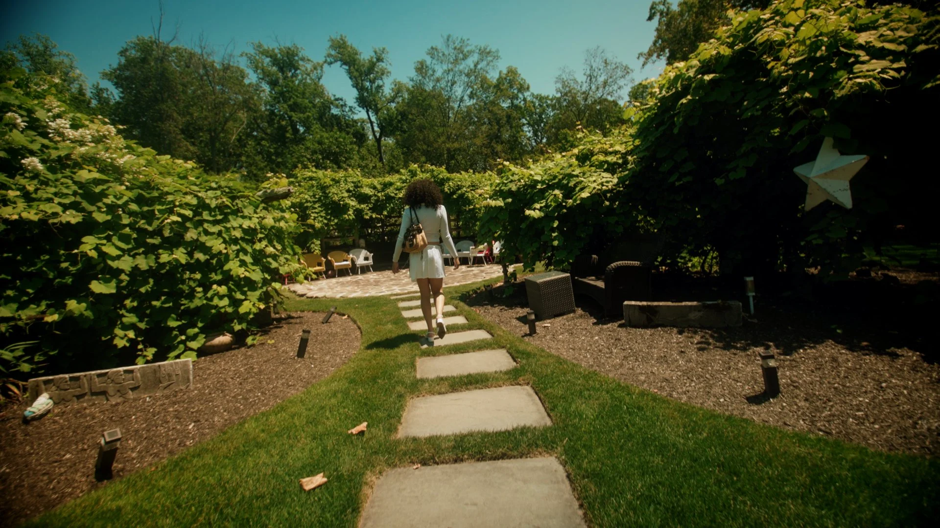 A woman walking on a stone pathway through a garden with lush green bushes and trees, and outdoor seating in the background under a blue sky.