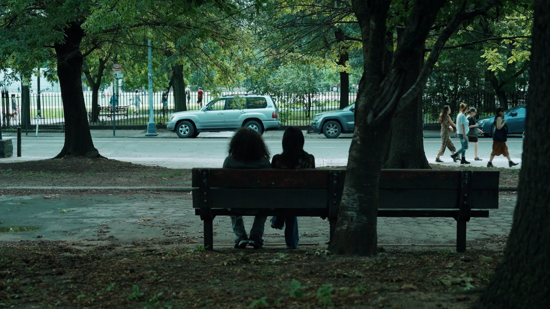 Two people sitting on a park bench under trees, looking toward a street with cars and pedestrians.