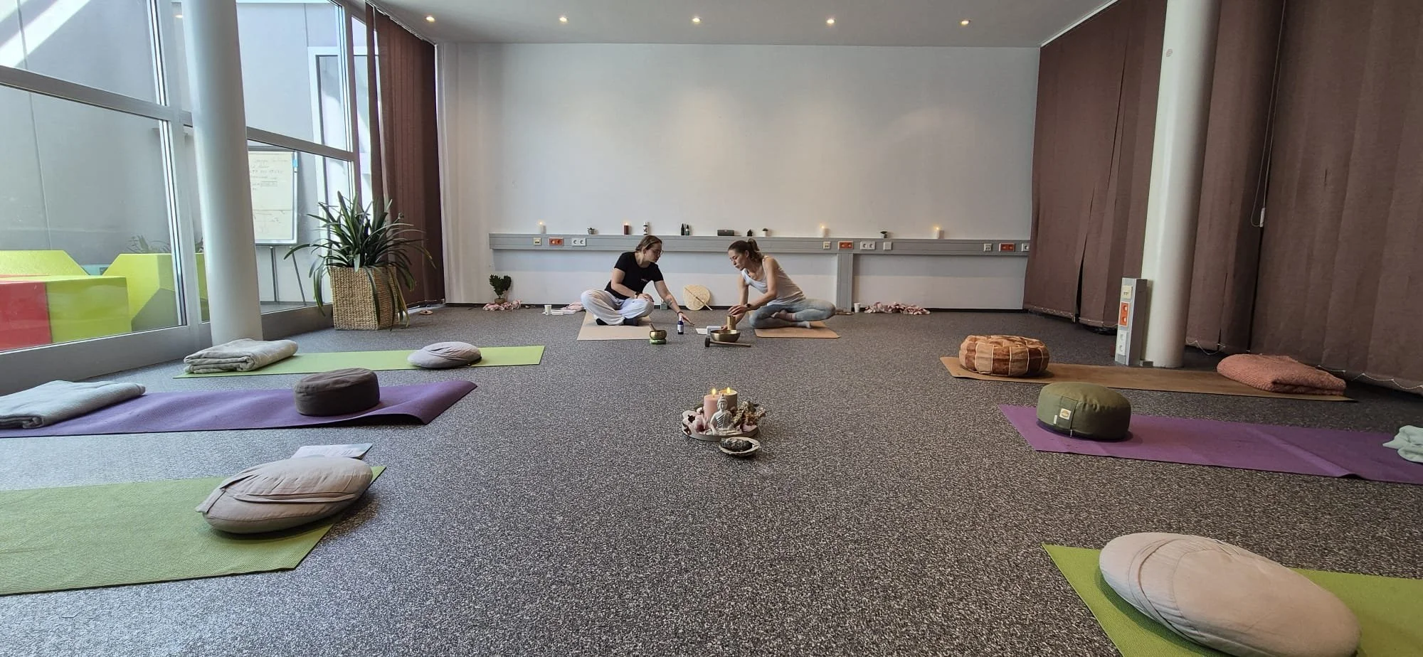 A yoga or meditation class setup in a spacious room with mats, cushions, and candles, two women preparing their mats near the wall, large windows with natural light, and a plant in the corner.