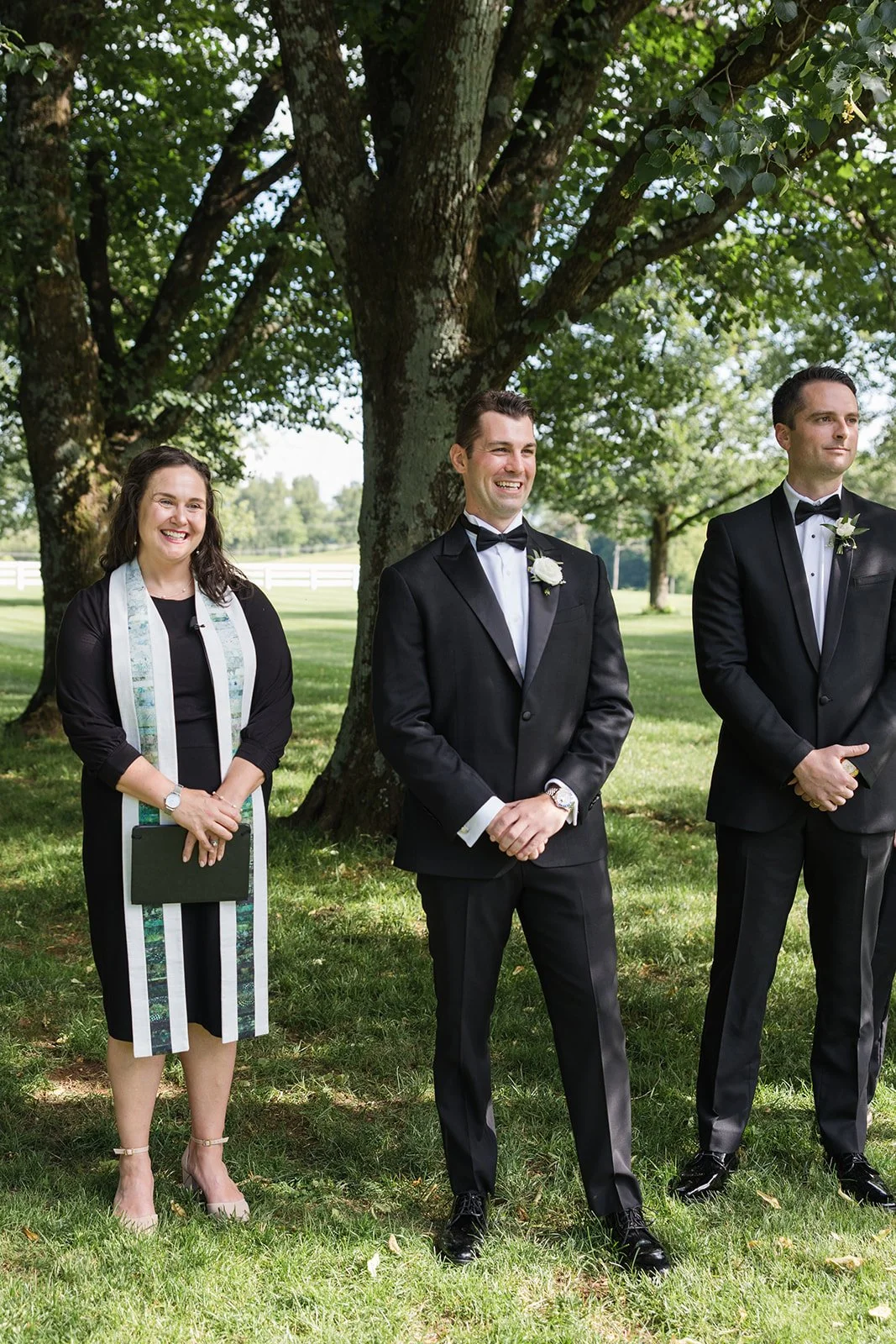 Three people standing outdoors under a large tree during a wedding ceremony, smiling and dressed formally. The woman on the left wears a black dress with a white and teal stole, the man in the middle wears a black tuxedo with a white shirt and black bow tie, and the man on the right also wears a black tuxedo with a white shirt and black bow tie.
