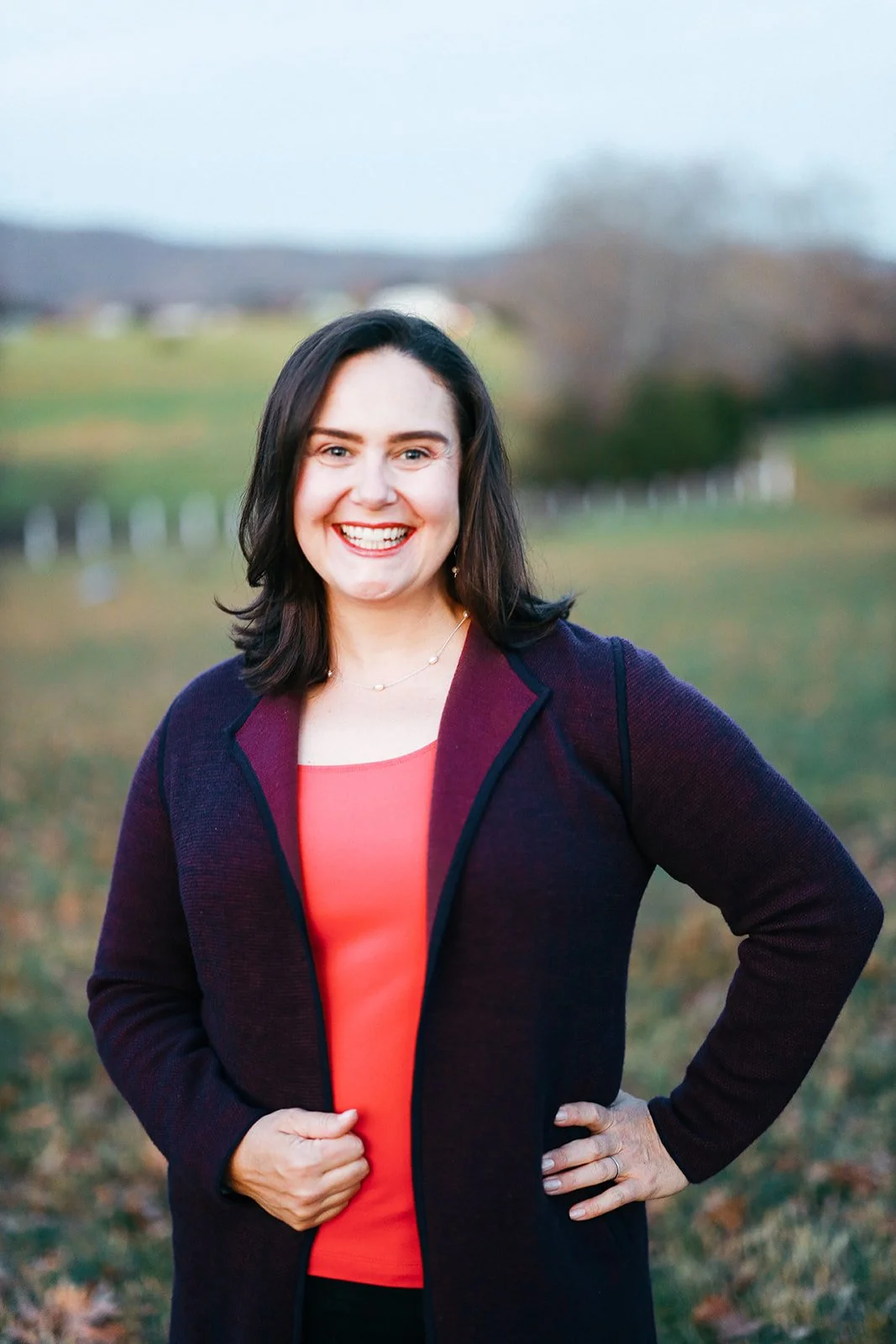 Kayla with dark brown hair and fair skin smiling at Grace Estate Winery with trees and a fence in the background. She is wearing a dark purple jacket over an orange top and has one hand on her hip.