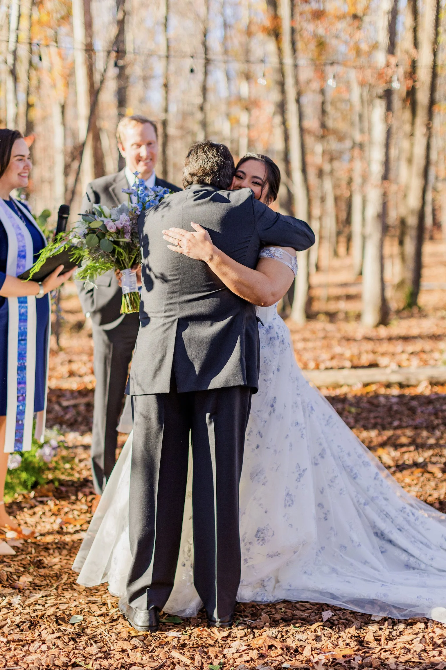 A couple exchanging vows and hugging during a wedding ceremony in a forest with autumn leaves, with a small group of people watching and smiling.