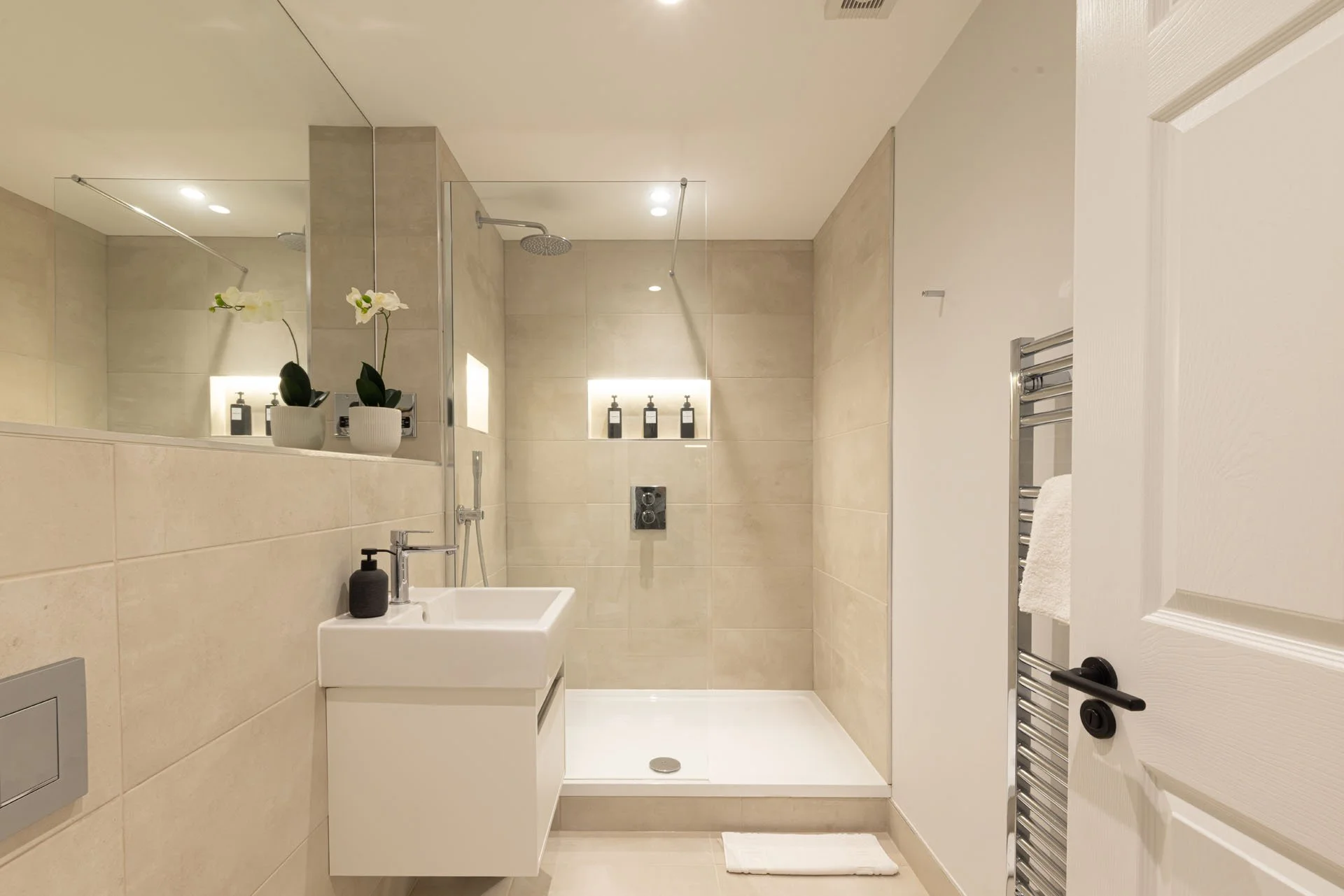 Modern bathroom with a walk-in shower, small white sink, and minimalistic design. There are two potted plants with white flowers on a ledge, a towel rack with a towel, and a large mirror on the wall.