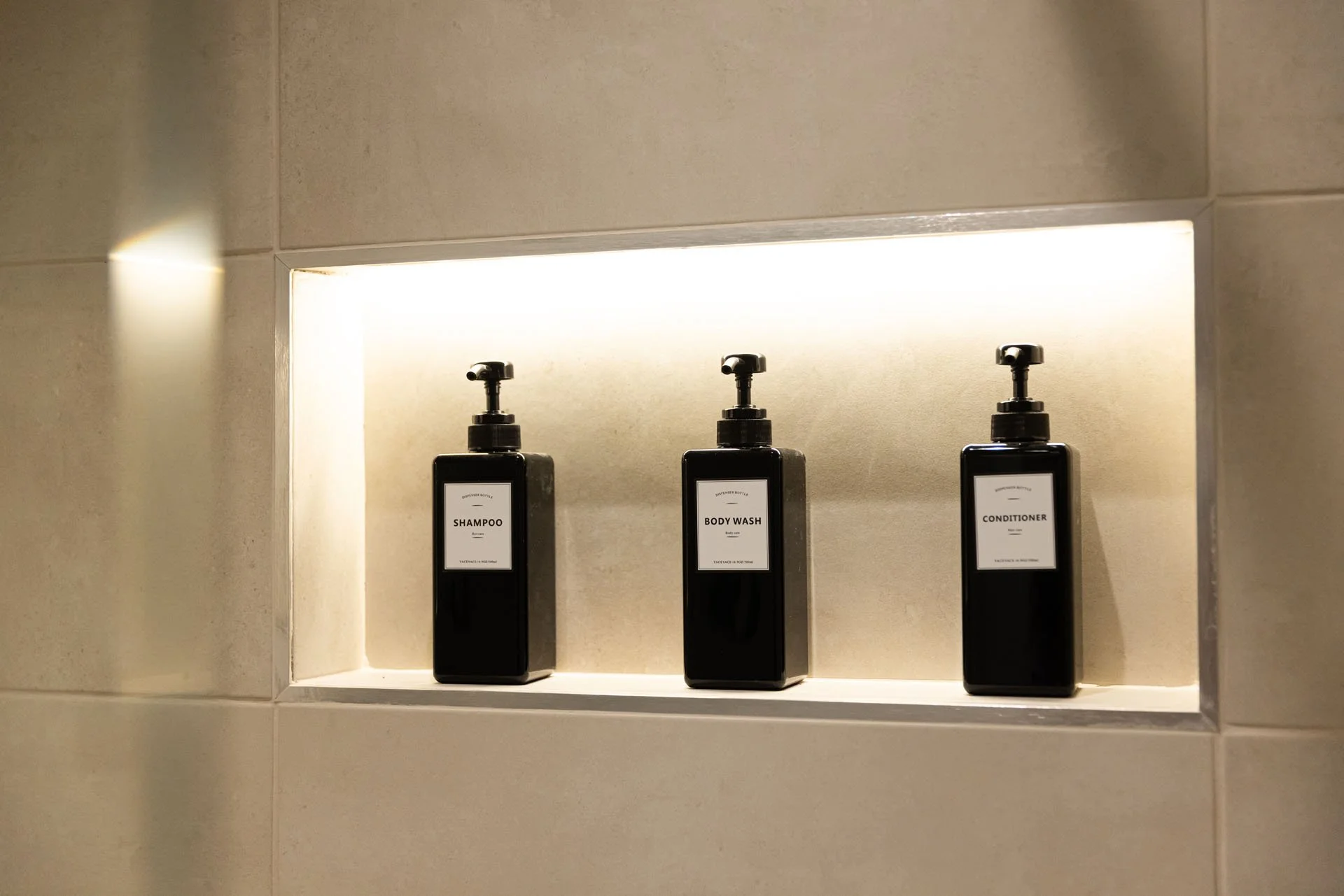 Three black bottles with pump dispensers labeled Shampoo, Body Wash, and Conditioner placed in a built-in wall shelf with backlighting.