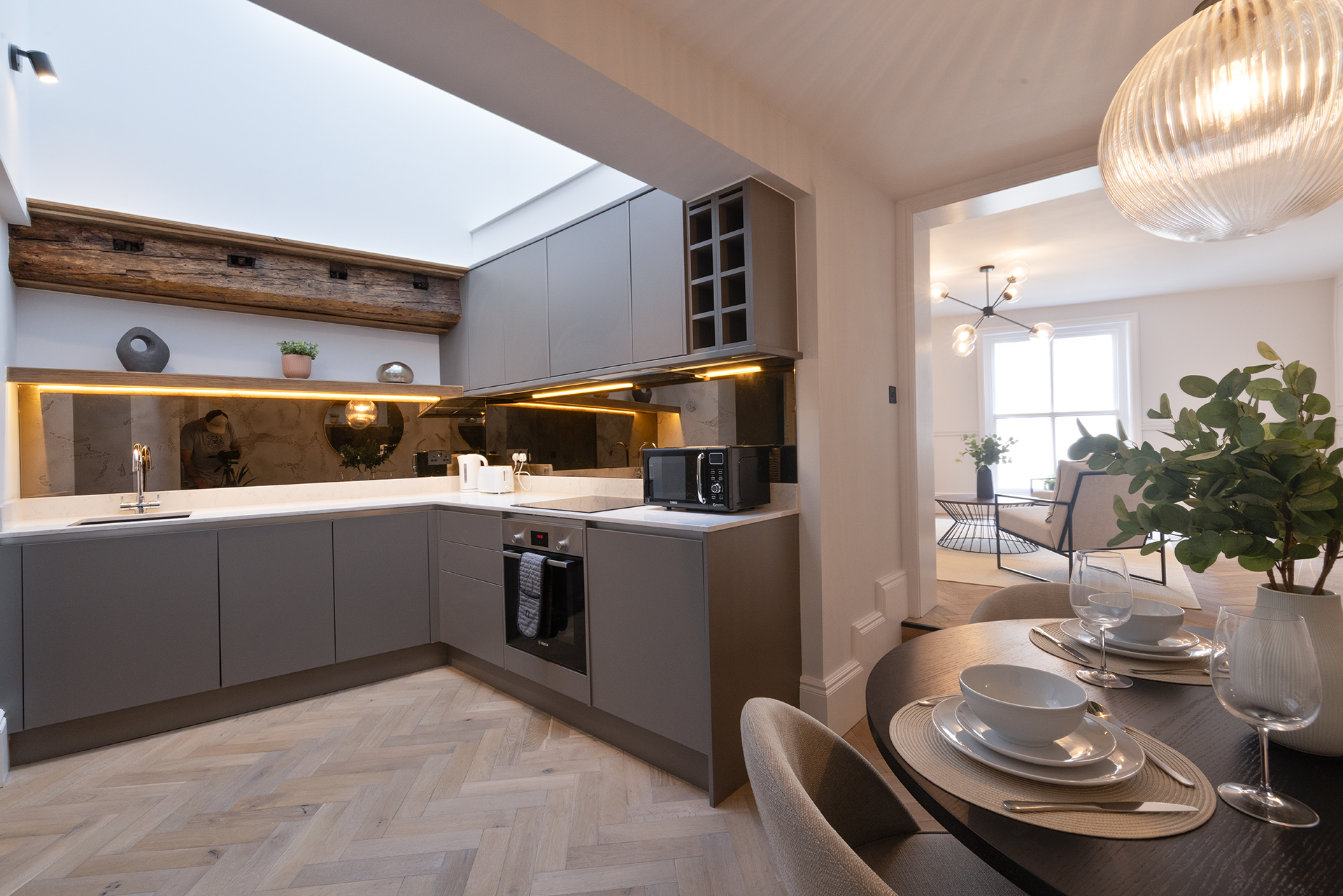 Modern kitchen with gray cabinets, white countertops, black backsplash, and wooden ceiling beam, adjacent to a dining area with table settings and a living room with large windows and contemporary lighting.