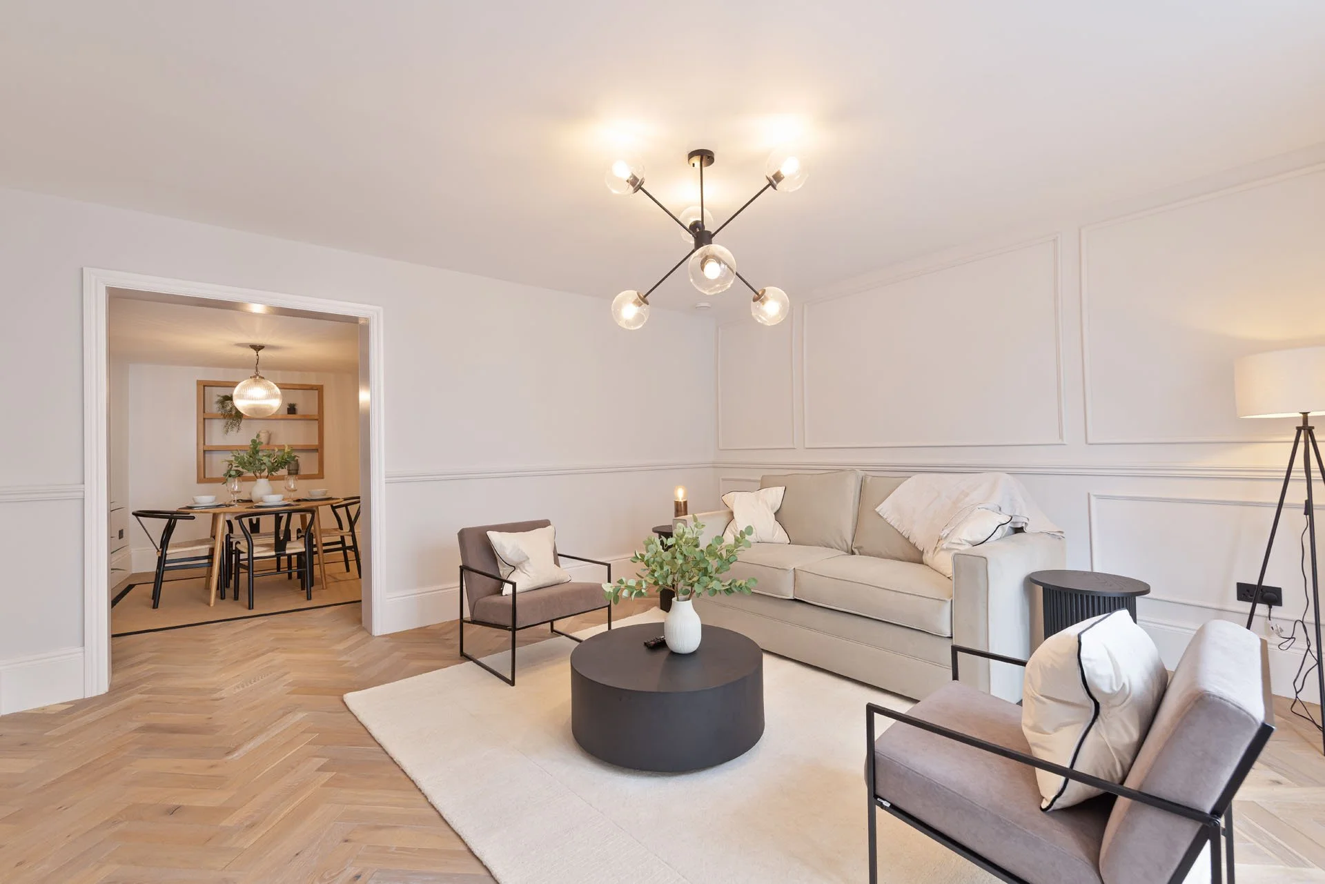 Modern living room with beige sofa, gray armchairs, black coffee table with a white vase and green foliage, wooden parquet flooring, white walls, and a black chandelier with spherical bulbs, connected to a dining area visible through a doorway.