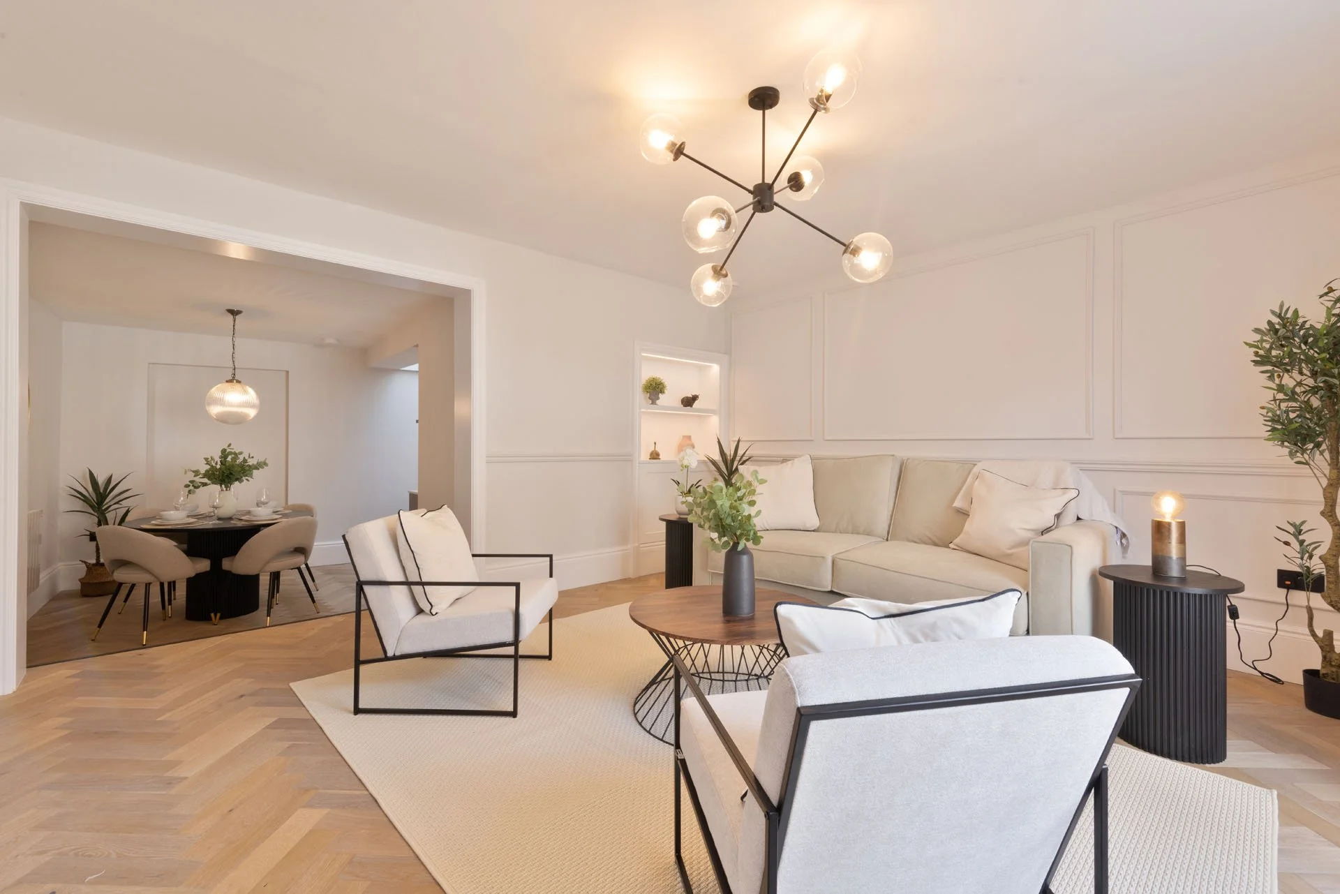 Bright living room with white walls, beige sofa, black side tables, wooden coffee table, modern chandelier, and potted plants, adjacent to a dining area with a round table and six chairs.