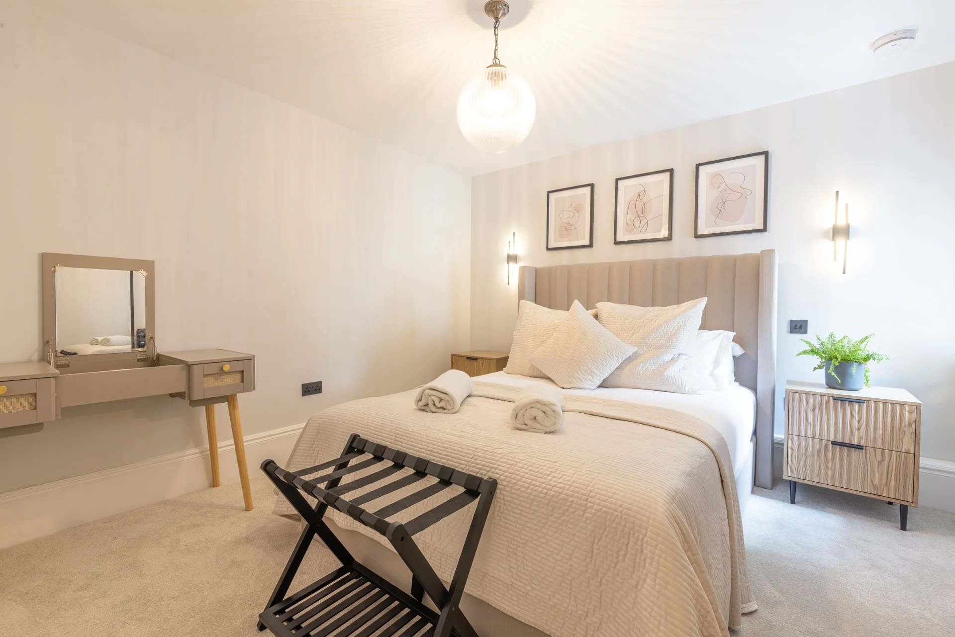 A neatly made bed with white linens and pillows in a modern bedroom, featuring a beige headboard, side tables, abstract artwork above the bed, a hanging light fixture, and a small vanity with a mirror.