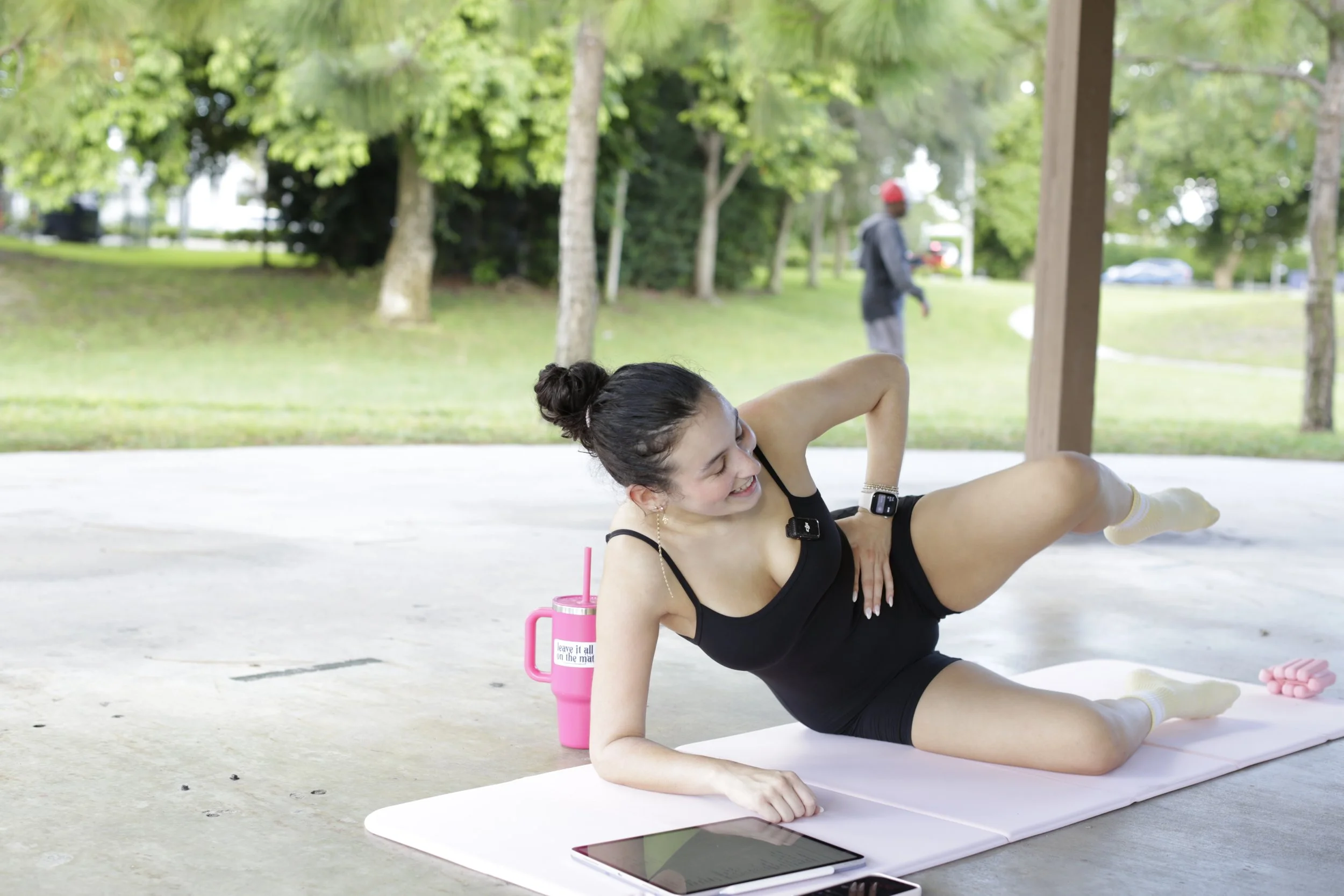 Young woman doing a side plank exercise outdoors, wearing black workout clothes and wristwatch, smiling, lying on a pink yoga mat, with a pink drink tumbler nearby, tablet and pink dumbbells on the ground, park background with trees and a person walking in the distance.