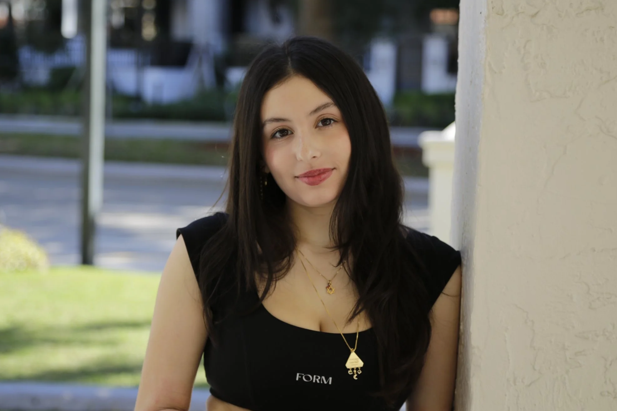 A young woman with long black hair, wearing a black top with the word 'FORM' on it, stands outdoors near a white wall, looking at the camera with a slight smile.