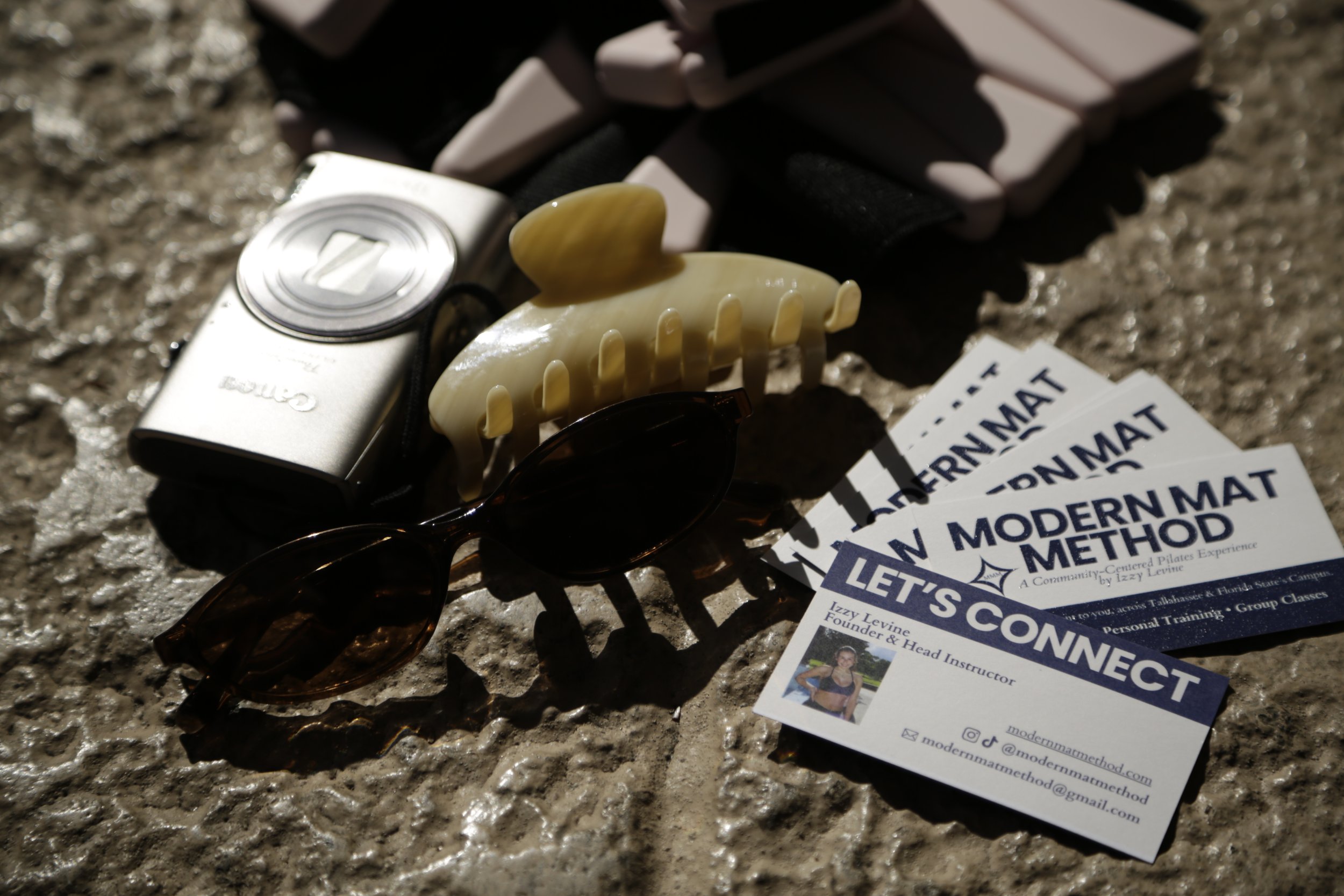 Sunlight illuminating various personal items on a textured surface, including a silver Canon camera, a cream-colored hair clip, a pair of dark sunglasses, a business card for Izzy Levine, and some printed informational sheets about a modern fitness method.