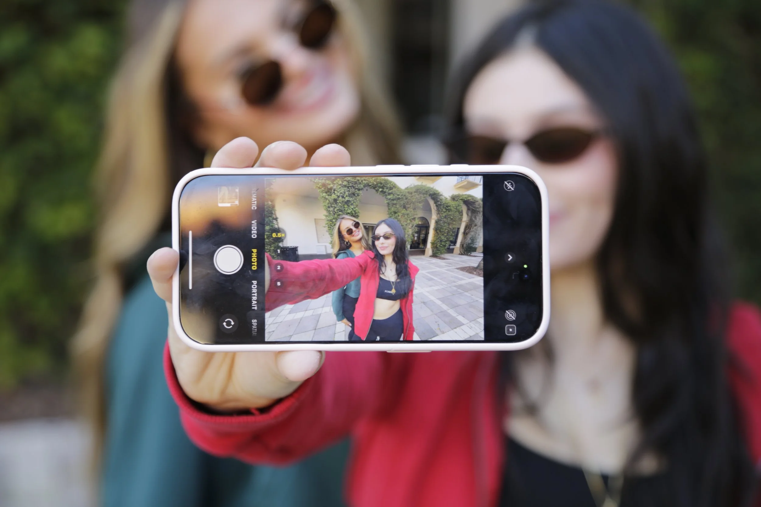 Two women taking a selfie with a smartphone, capturing their faces and backgrounds, both wearing sunglasses and smiling.